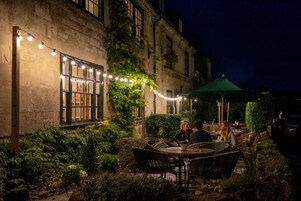 An outdoor seating area with tables and chairs, surrounded by greenery and a building facade.