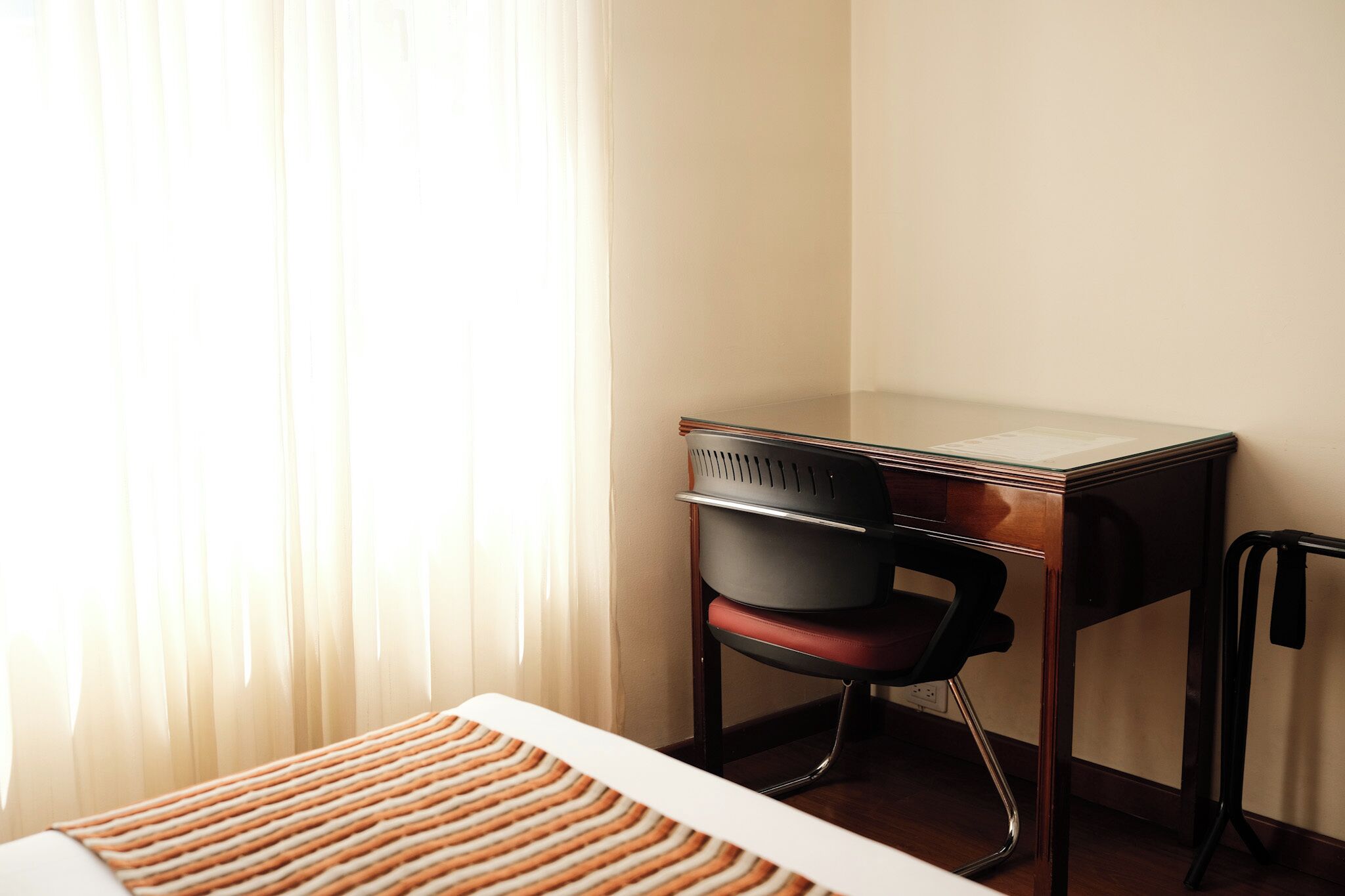 Habitación de hotel con escritorio de madera, una silla de oficina roja, cama blanca con colcha a rayas y cortinas traslúcidas que dejan entrar luz natural.