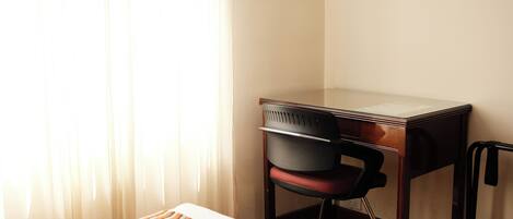 Habitación de hotel con escritorio de madera, una silla de oficina roja, cama blanca con colcha a rayas y cortinas traslúcidas que dejan entrar luz natural.