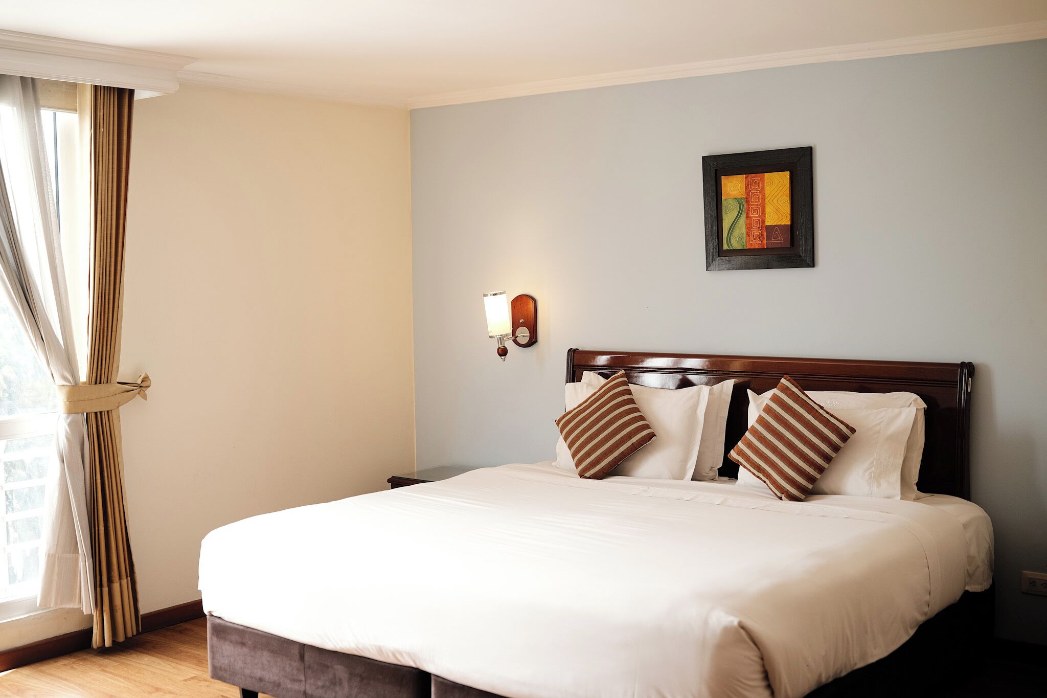 Un lit bien fait, avec du linge de lit blanc et deux coussins à rayures, une tête de lit en bois, un luminaire fixé au mur et une œuvre d’art encadrée accrochée au mur.