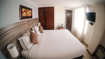 Quarto de hotel com uma cama grande, cabeceira de madeira, mesa de cabeceira com abajur, televisão na parede e porta com cortinas.
