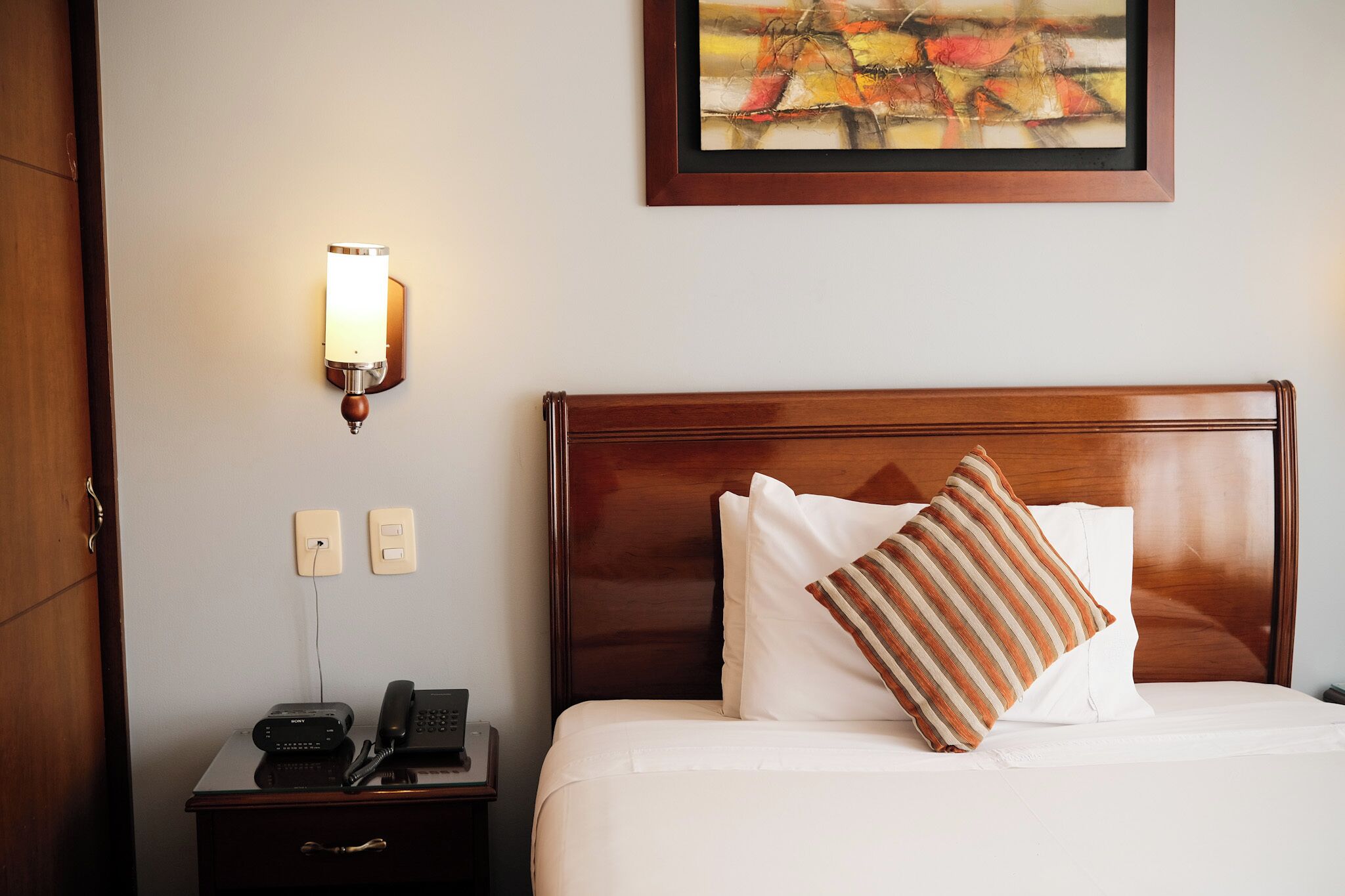 Habitación de hotel con cabecera de madera, mesita de noche con teléfono e iluminación en la pared.