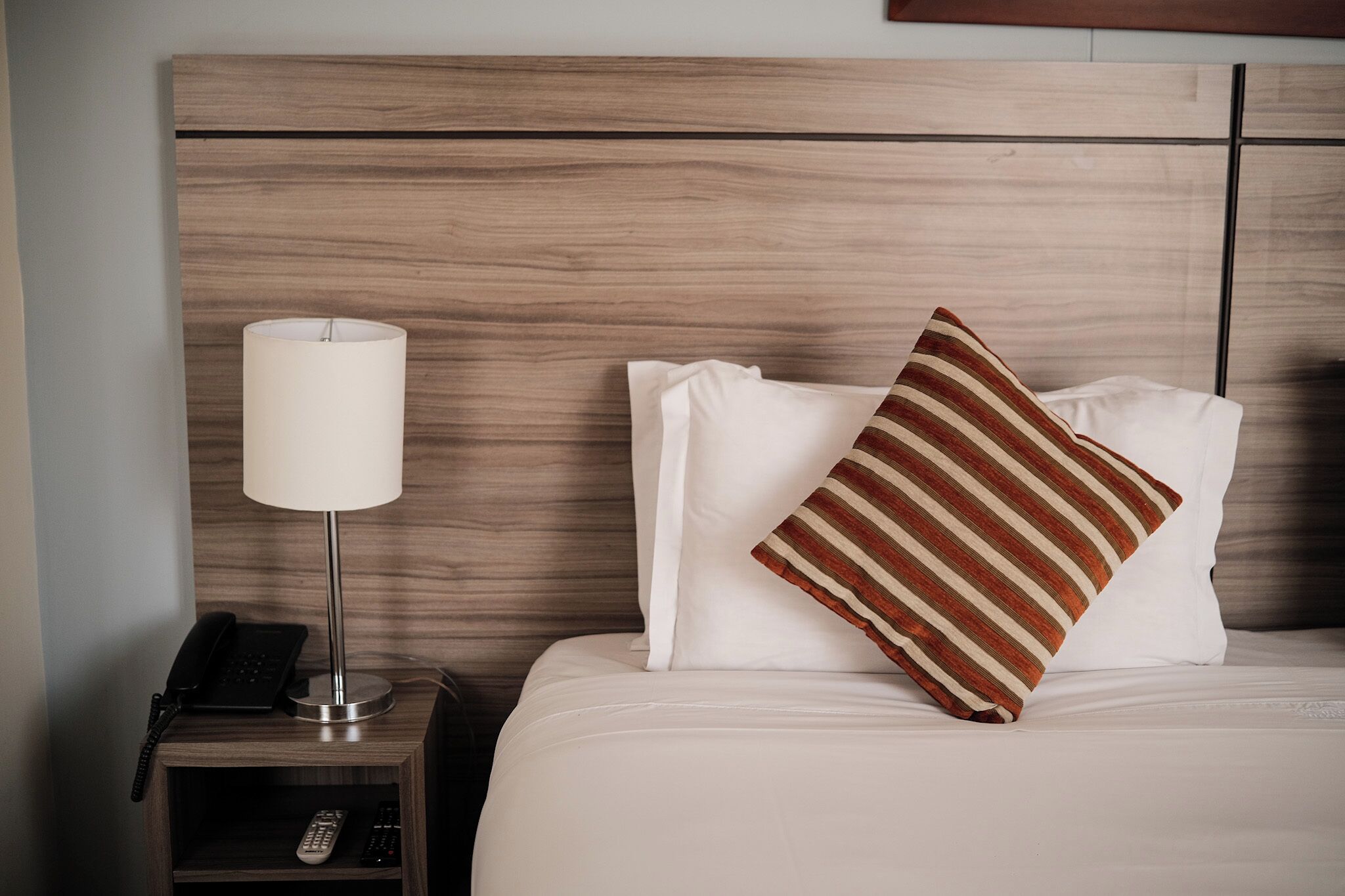 Un lit bien fait, avec du linge de lit blanc, un oreiller à rayures, une lampe de chevet et un téléphone posés sur une table de chevet en bois.