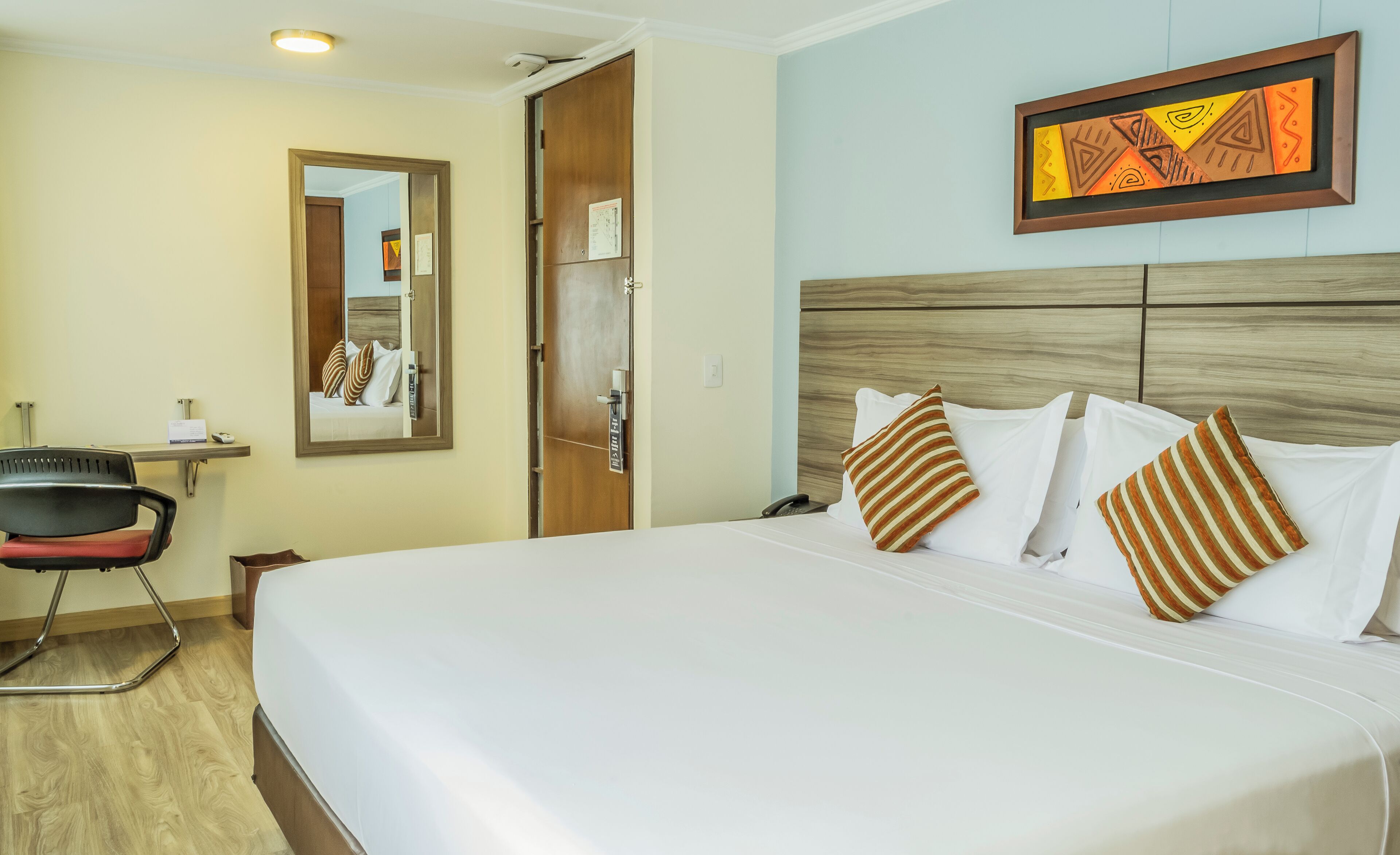 Une chambre d’hôtel avec un lit, un bureau, une chaise, un miroir et un tableau encadré au mur.