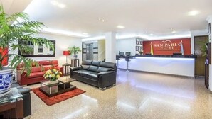 Un hall de hotel con mostrador de recepción, un sofá rojo, un sofá de cuero negro y una mesa de centro con flores.