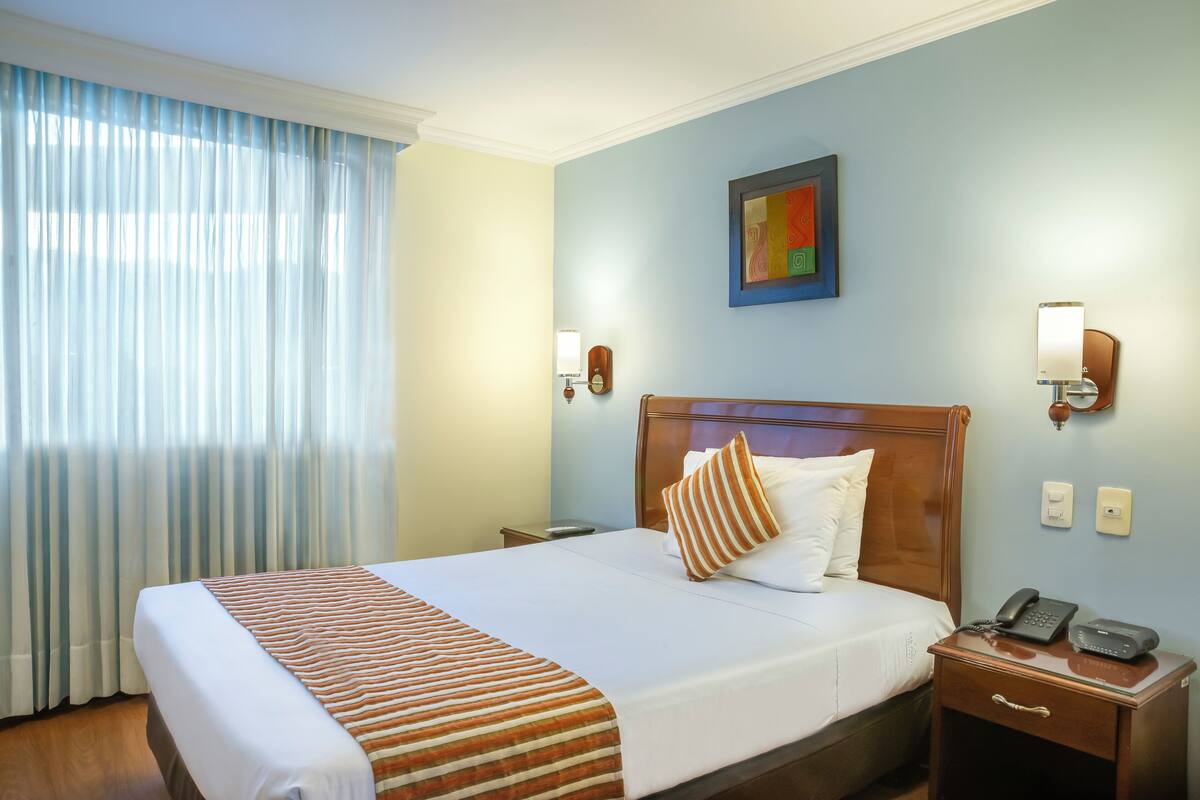 Una habitación de hotel con una cama grande, un cabecero de madera, una mesita de noche con un teléfono y una ventana con cortinas.