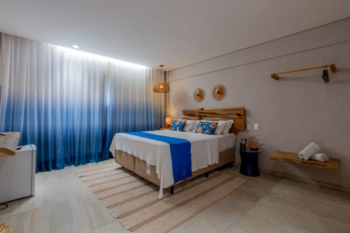 A modern bedroom with a large bed, a bedside table, a nightstand, a hanging light fixture, and a view of the outdoors through sheer curtains.