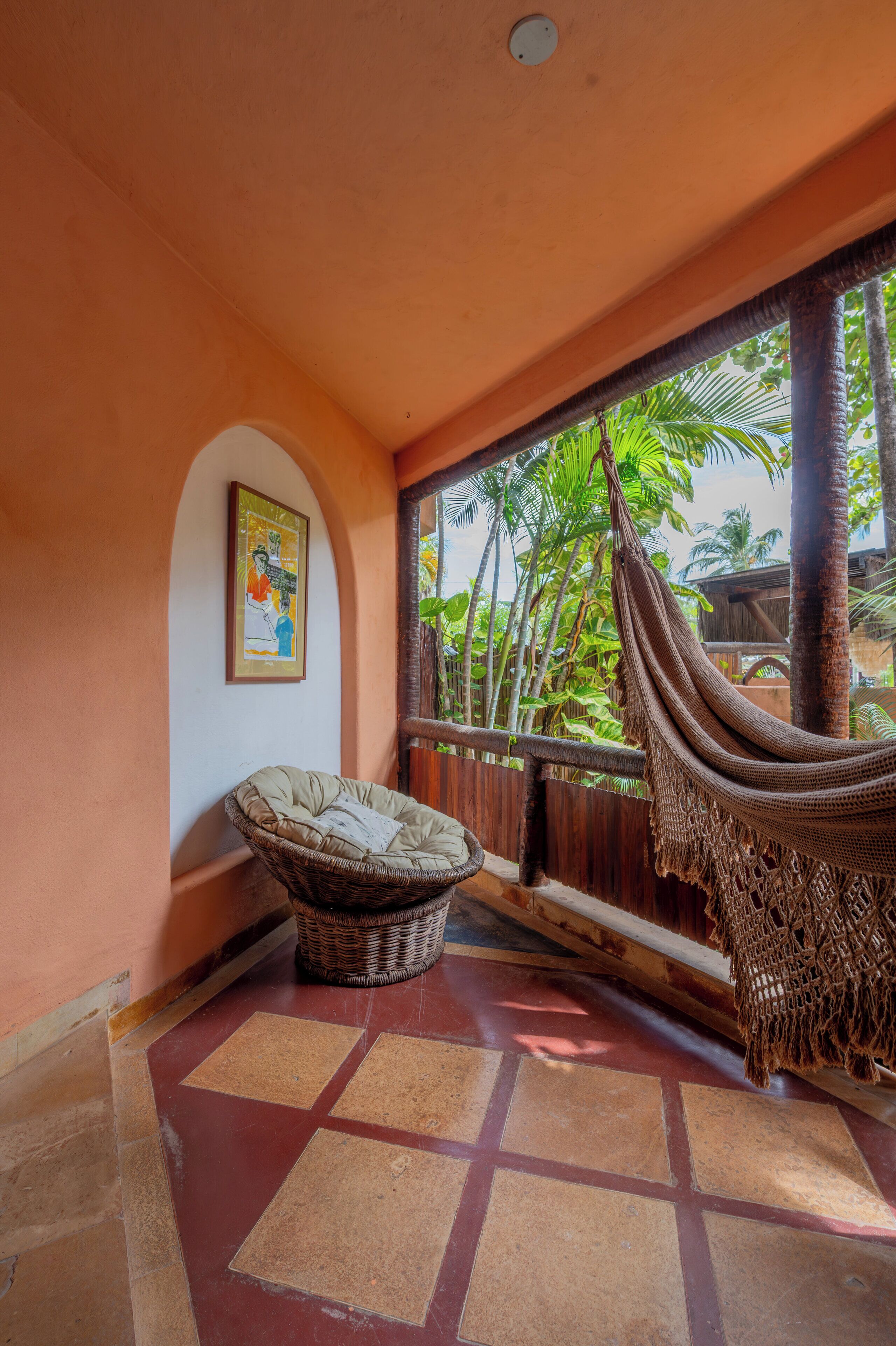 A room with a hammock, a wicker chair, a framed picture on the wall, and a view of palm trees through a window.