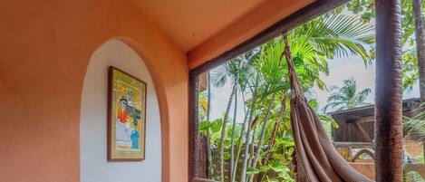 A room with a hammock, a wicker chair, a framed picture on the wall, and a view of palm trees through a window.
