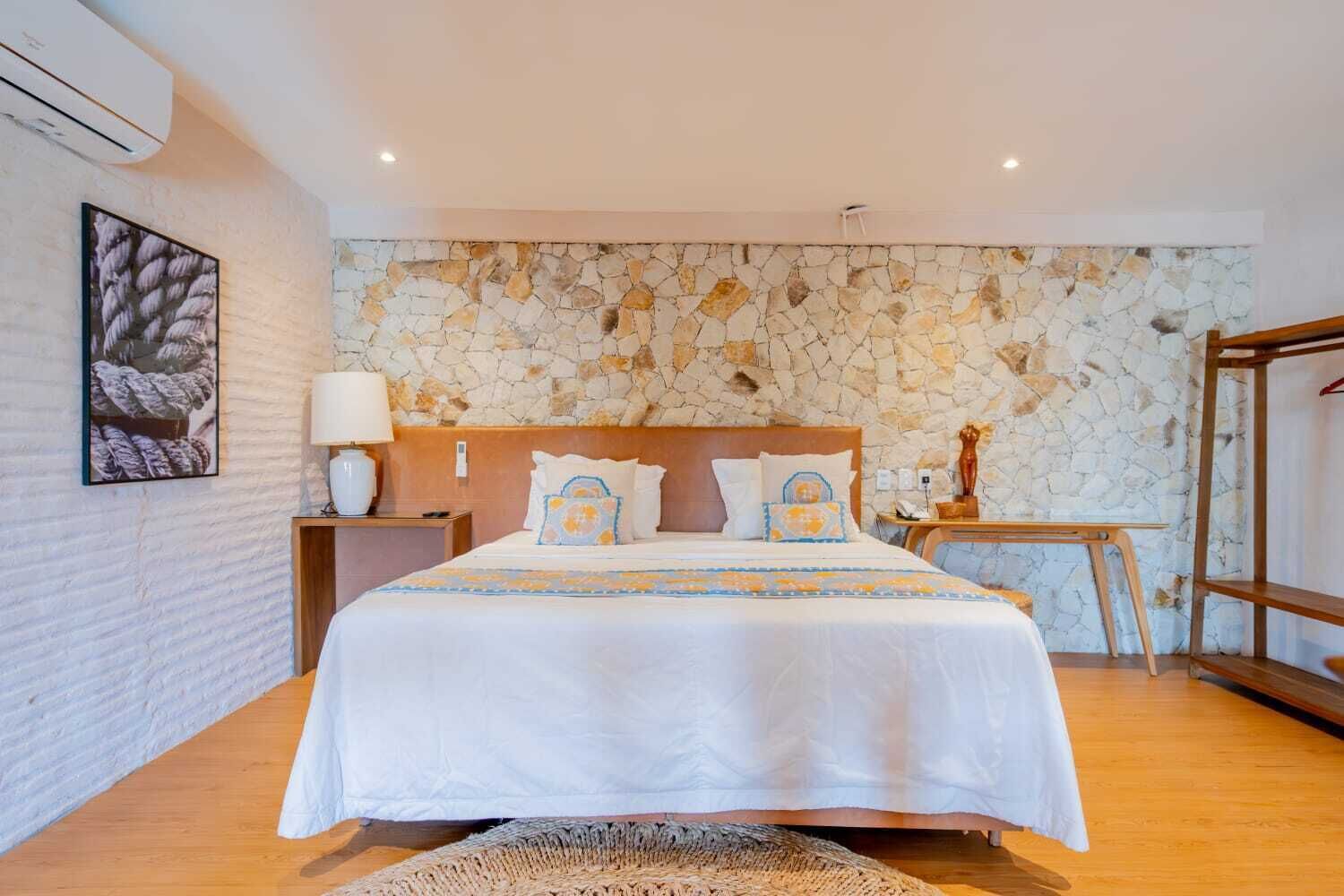 Une chambre avec un mur en pierre, un lit avec une literie blanche, une table de chevet, une lampe et une petite table.