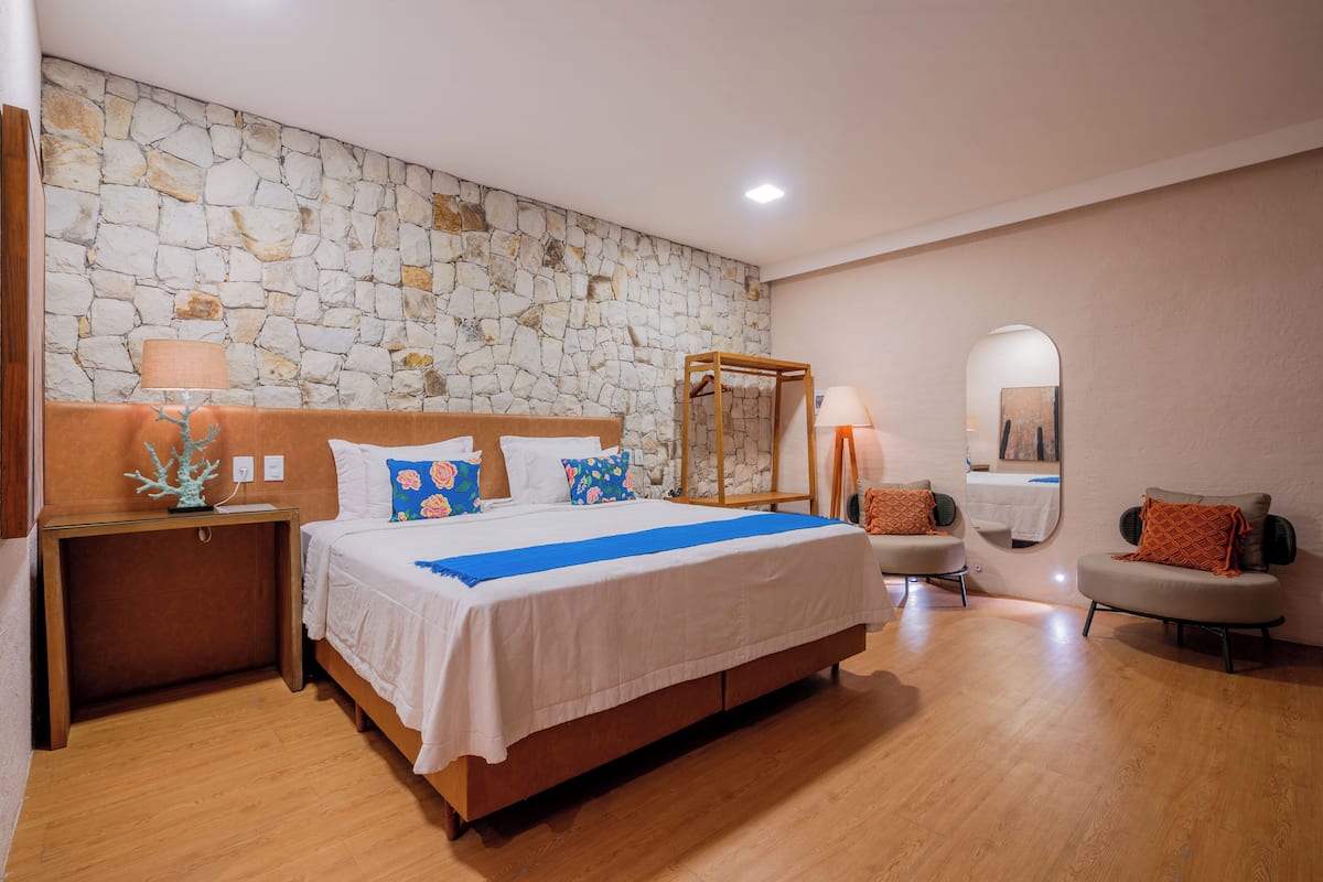 A bedroom with a stone wall, a bed with white bedding and colorful pillows, a wooden nightstand, a chair, and a mirror.