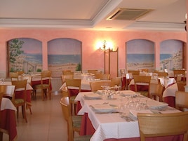 Un comedor con mesas dispuestas para el servicio, sillas y vista al mar a través de ventanas arqueadas.