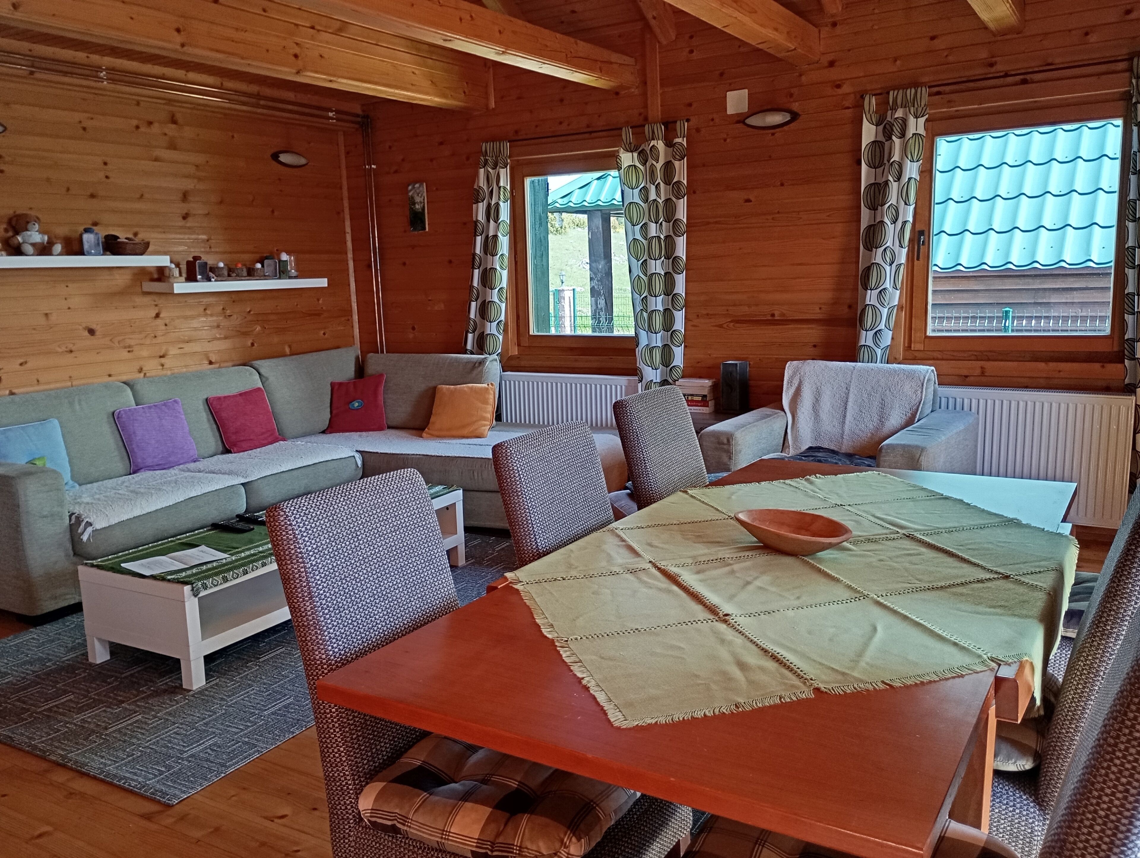 A cozy cabin interior with a wooden interior, a dining table with a bowl, a sofa with cushions, and a window with patterned curtains.
