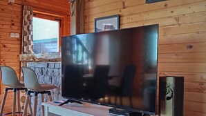 Interior de madera con una televisión sobre un soporte blanco, paredes de madera y una ventana con cortinas.