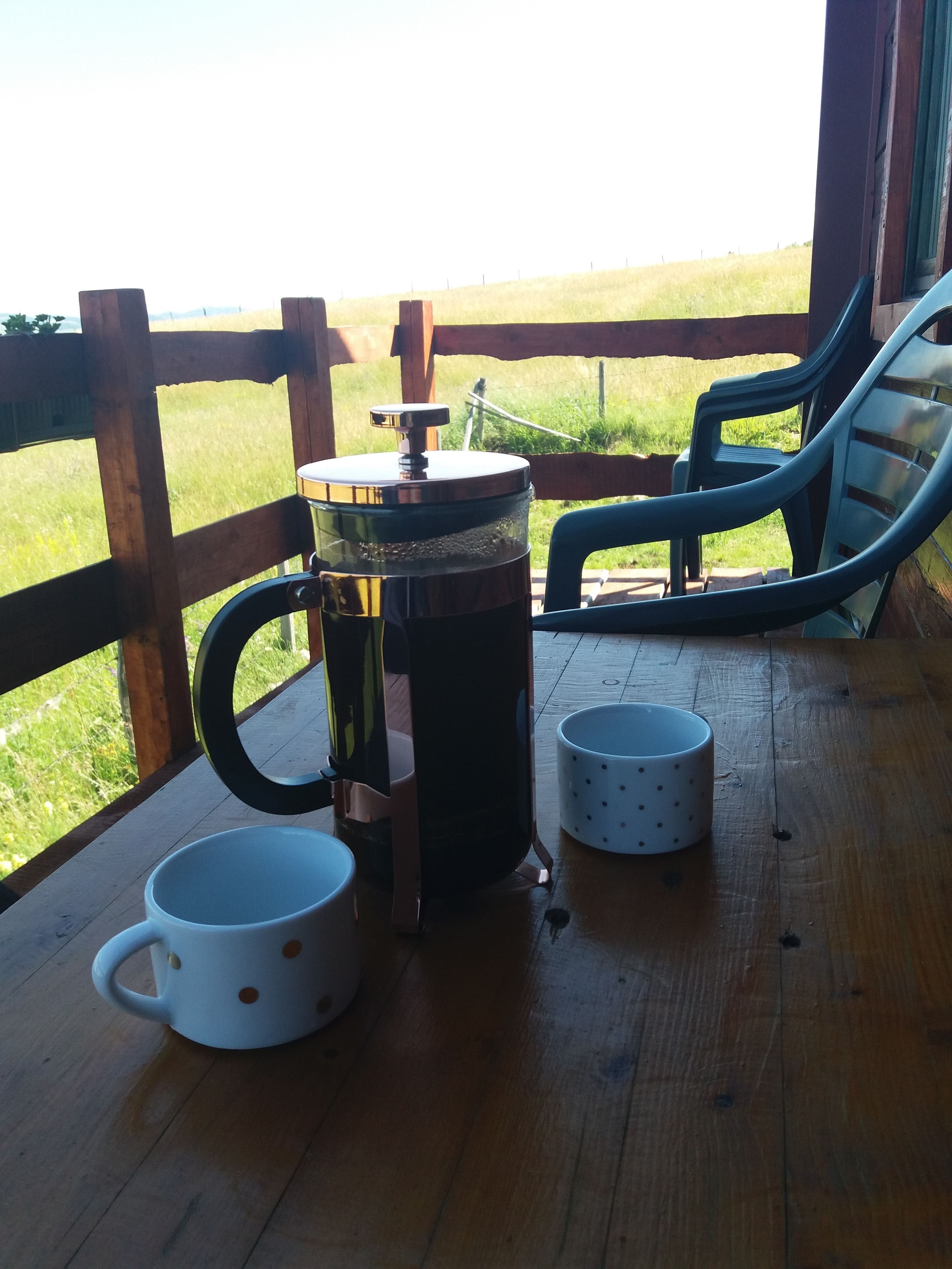 A wooden deck with a French press, two white mugs with polka dots, and a blue chair.