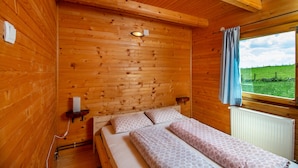 Un dormitorio de cabaña de madera con una cama, una mesita de noche, un ventanal con vista a un campo verde y una lámpara de techo.
