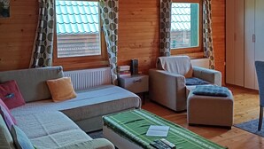 Interior acogedor de una cabaña de madera con sofá, sillón y reposapiés, vigas de madera en el techo y vistas a través de las ventanas.