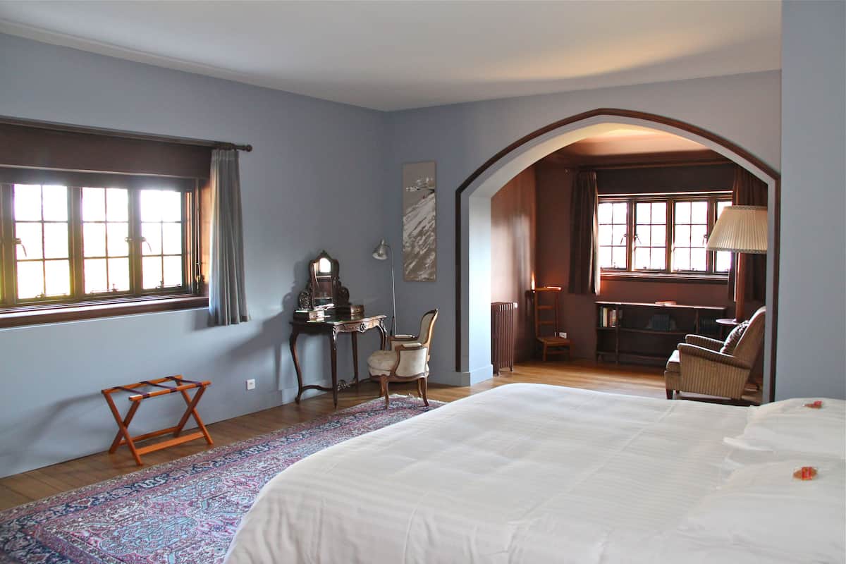 A bedroom with a large bed, a desk, a chair, and a view of a kitchen through an archway.