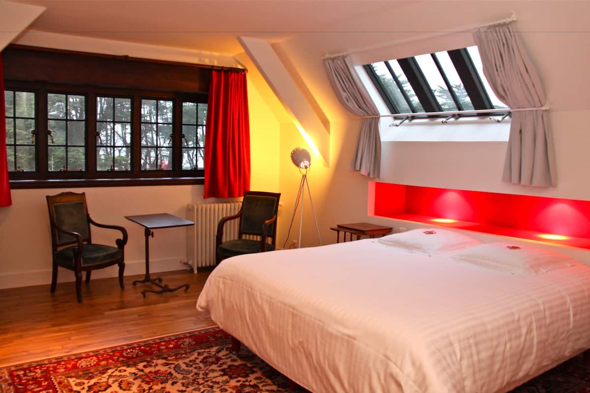 A bedroom with a large bed, two chairs, a small table, a floor lamp, and a window with curtains.