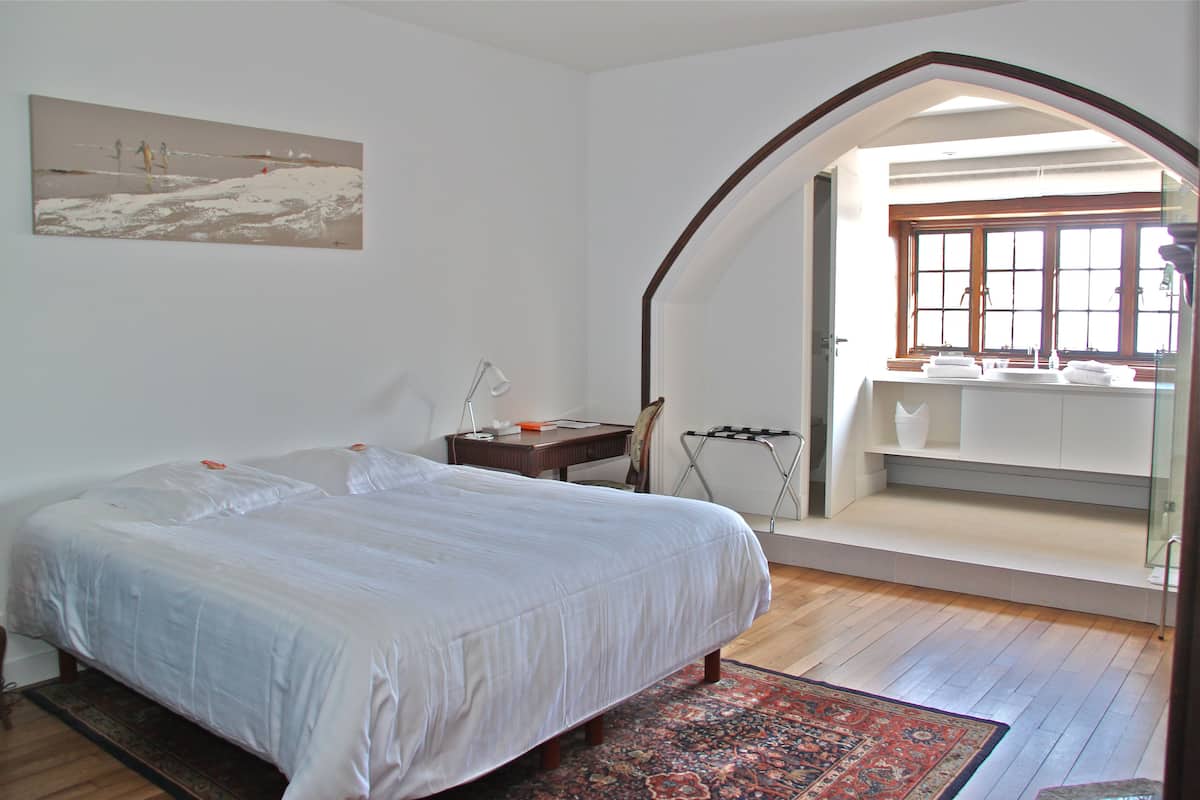 A bedroom with a bed, a desk, a chair, a window, and a bathroom visible through an archway.