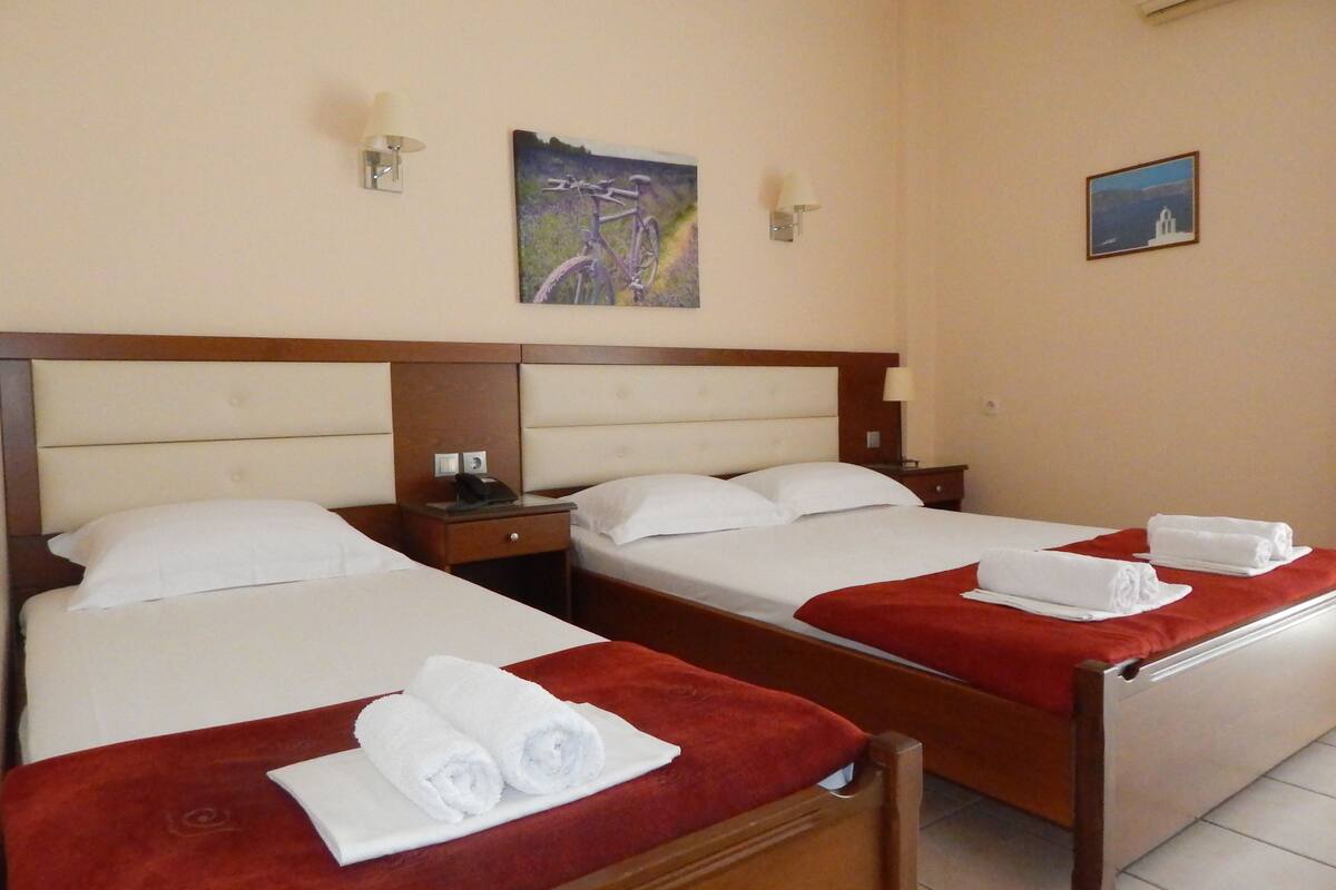 Habitación de hotel con dos camas, cada una con una manta roja y toallas enrolladas. Hay dos mesitas de noche con lámparas y un cuadro en la pared.