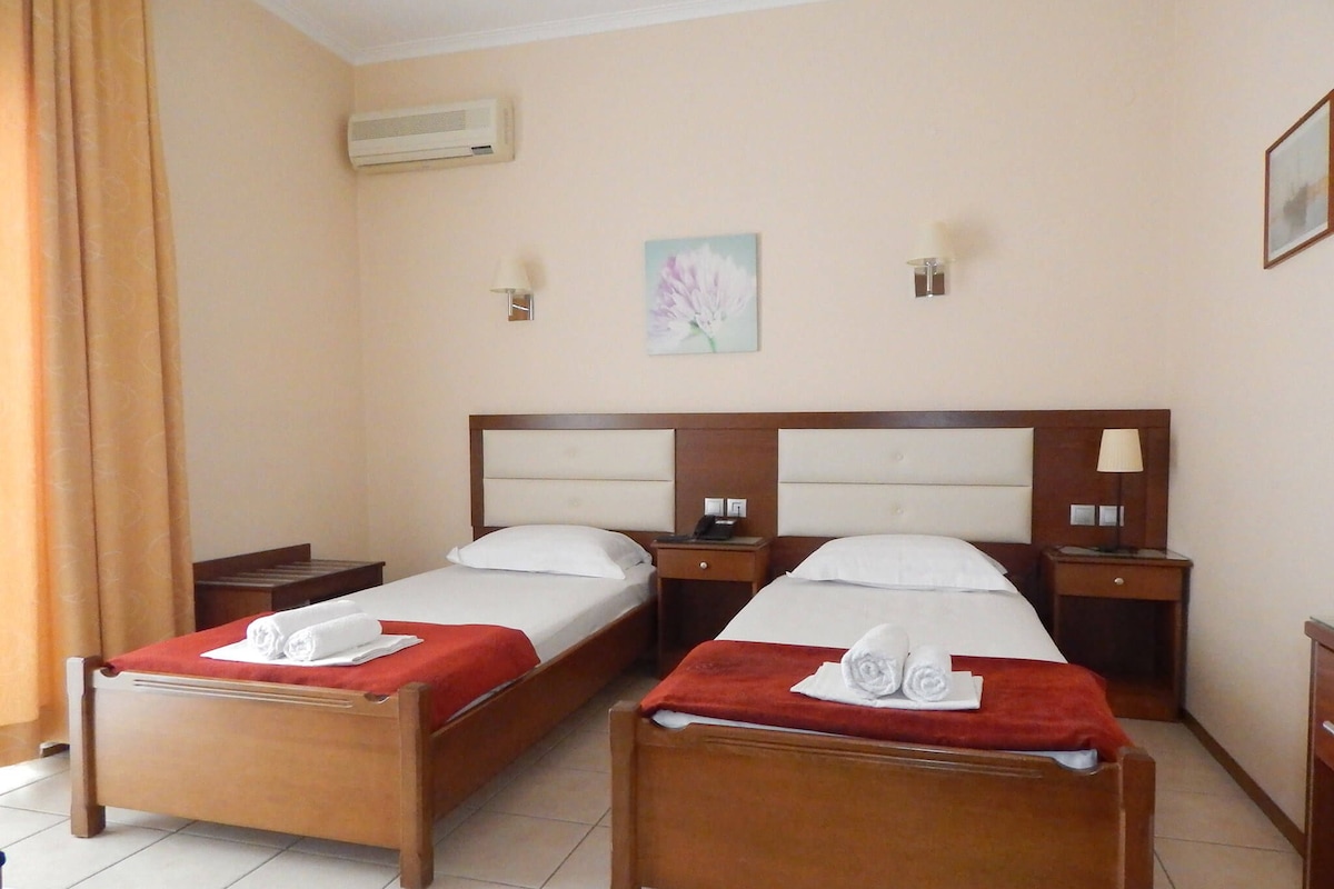 Una habitación de hotel con dos camas individuales, cada una con estructura de madera, ropa blanca y colchas rojas. Hay mesitas de noche con lámparas, un aire acondicionado de pared y un cuadro enmarcado en la pared.