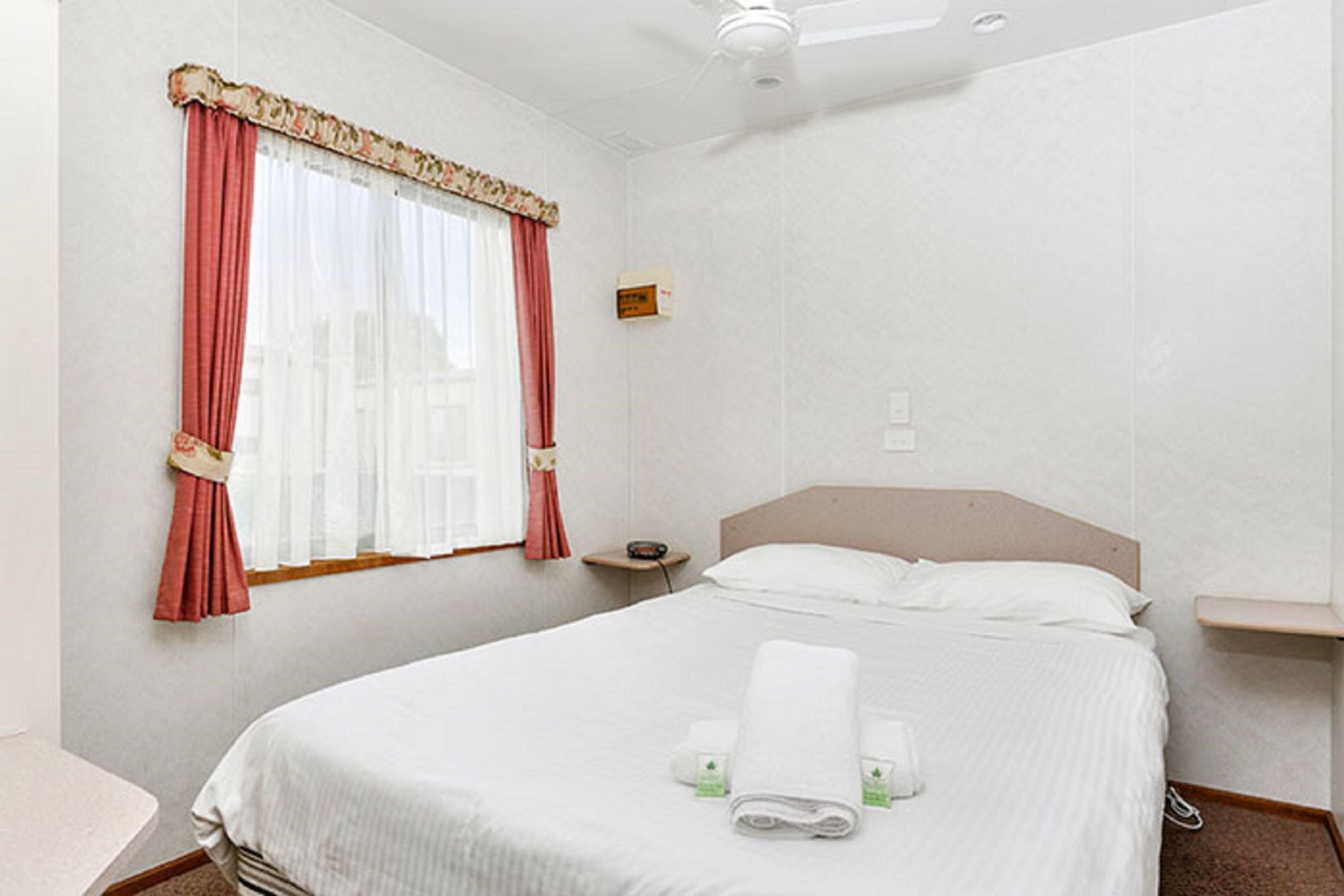 Ein Hotelzimmer mit Bett, Nachttisch, einem Fenster mit Vorhängen und einem Deckenventilator.