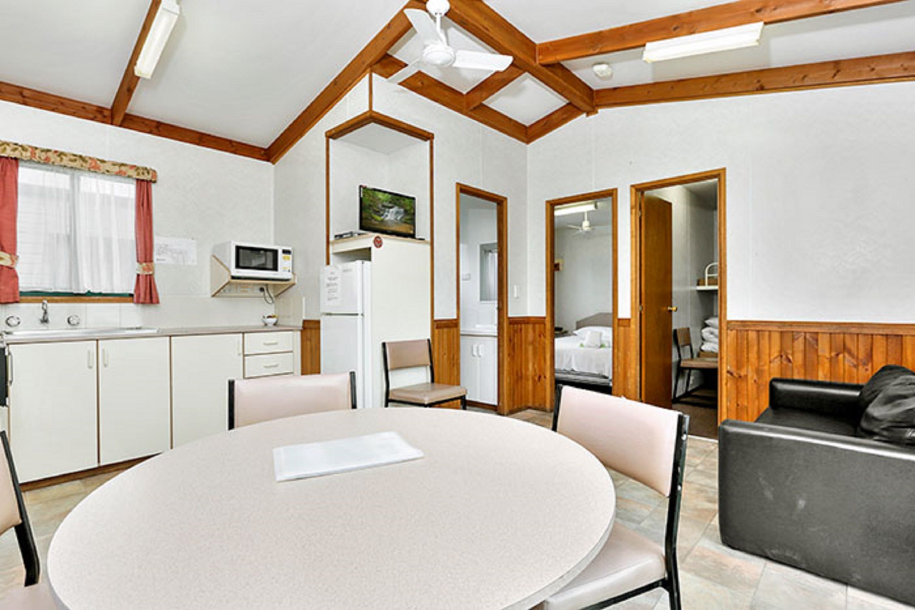 A compact room with a round table, chairs, a microwave, a refrigerator, and a bed with a wooden headboard.