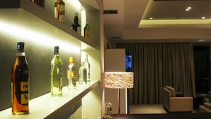 Un moderno hall de hotel con una zona de bar que exhibe varias botellas de licor, un espacio de descanso con televisor y un amplio ventanal con cortinas.