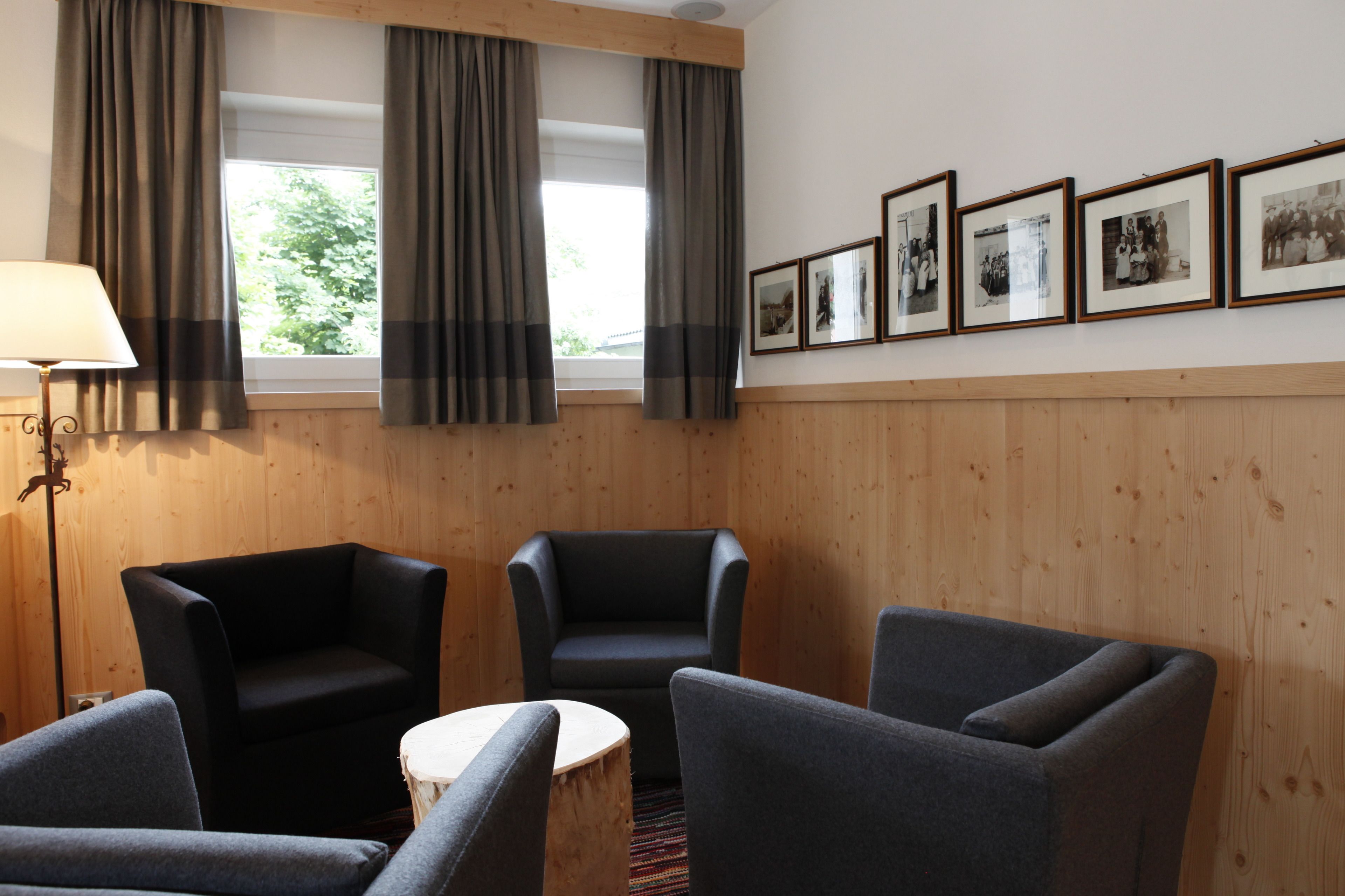 Ein Zimmer mit Holzvertäfelung, zwei grauen Sesseln, einem runden Holztisch und einer Stehlampe. An der Wand hängen vier gerahmte Fotografien.