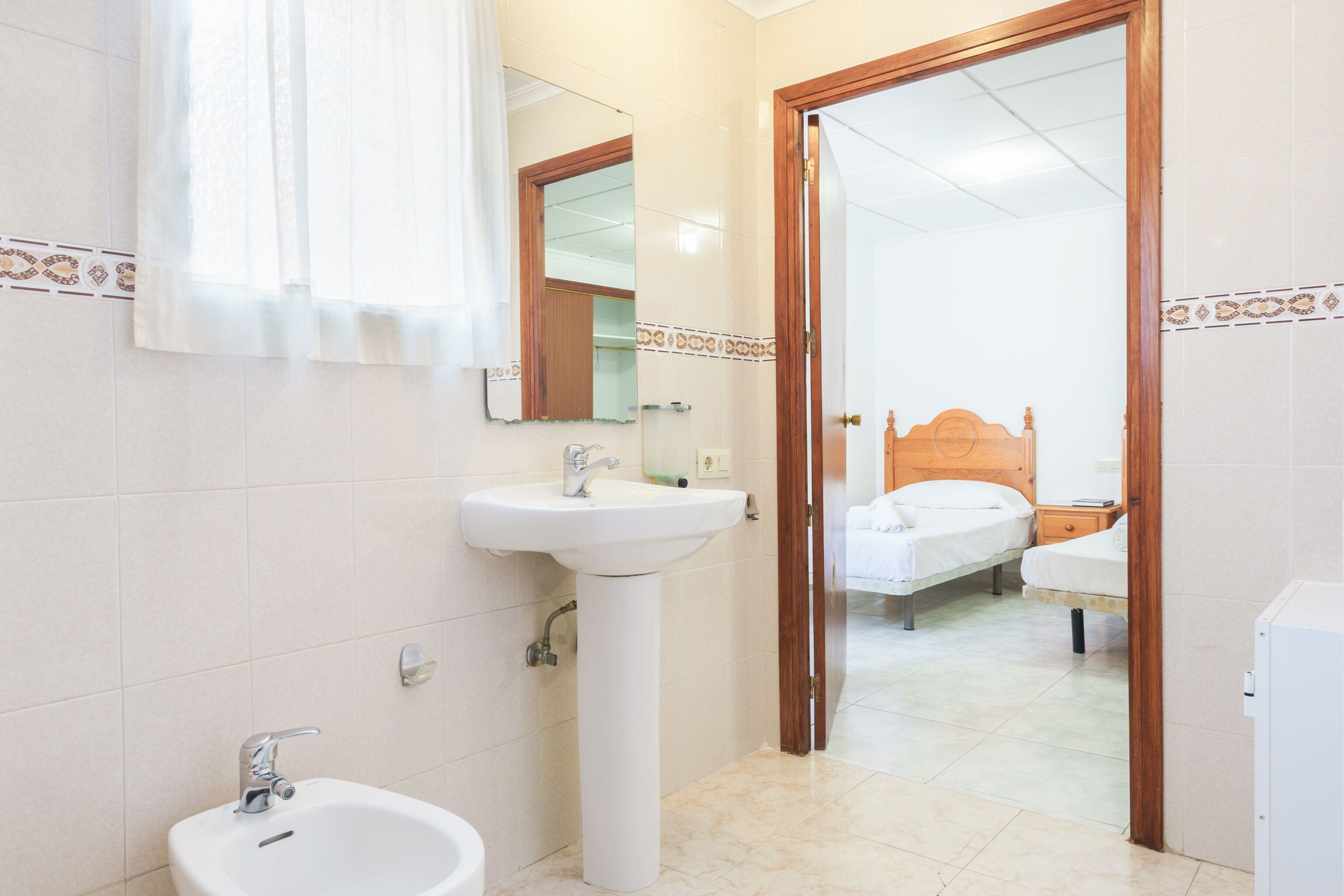 A bathroom with a sink, mirror, and a door leading to a bedroom with a bed and bedside table.
