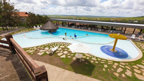 A large outdoor swimming pool with a thatched roof structure, surrounded by lounge chairs and umbrellas, with a view of the landscape in the background. - Fazenda Monte Castelo (Sairé)