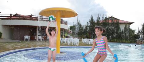 A swimming pool area with a water play structure, surrounded by white chairs and tables, and a building with a red roof in the background.