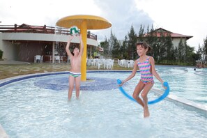 Zona de piscina con estructura de juegos acuáticos, rodeada de sillas y mesas blancas, y un edificio con tejado rojo al fondo.