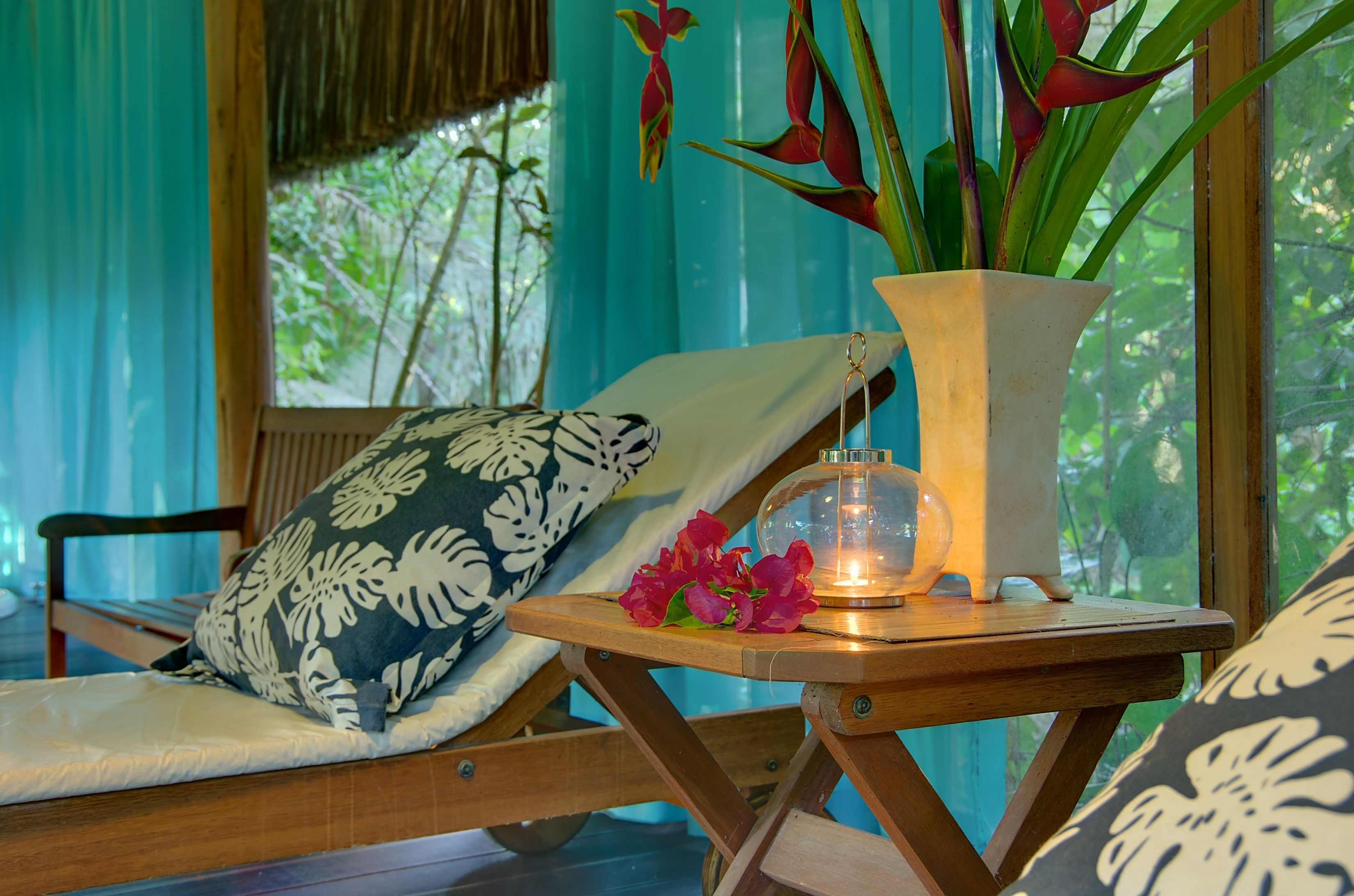 A wooden lounge chair with a patterned pillow, a small table with a vase of flowers, and a candle.