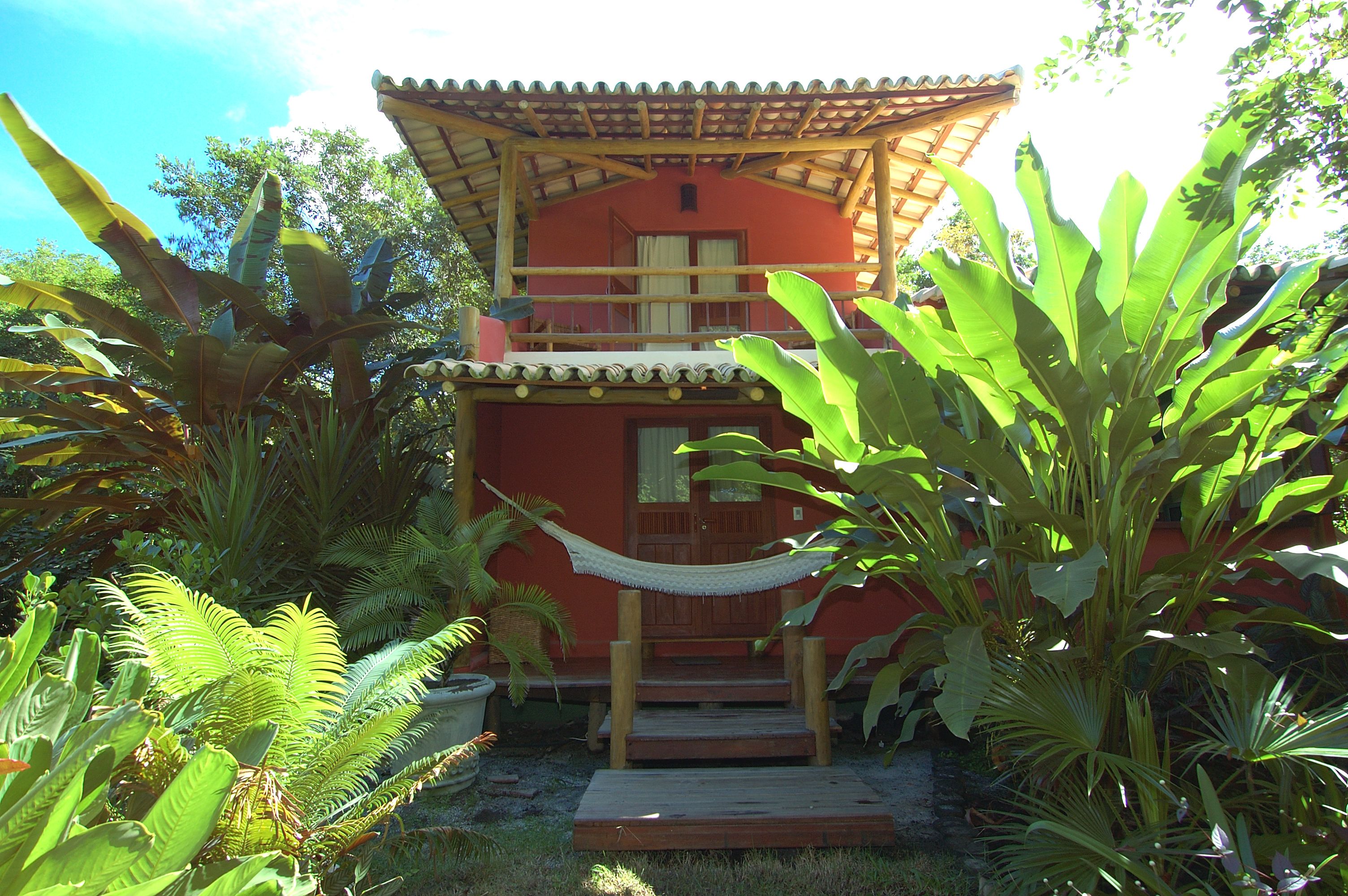 A two-story building with a red facade, a wooden balcony, and a hammock in the garden.