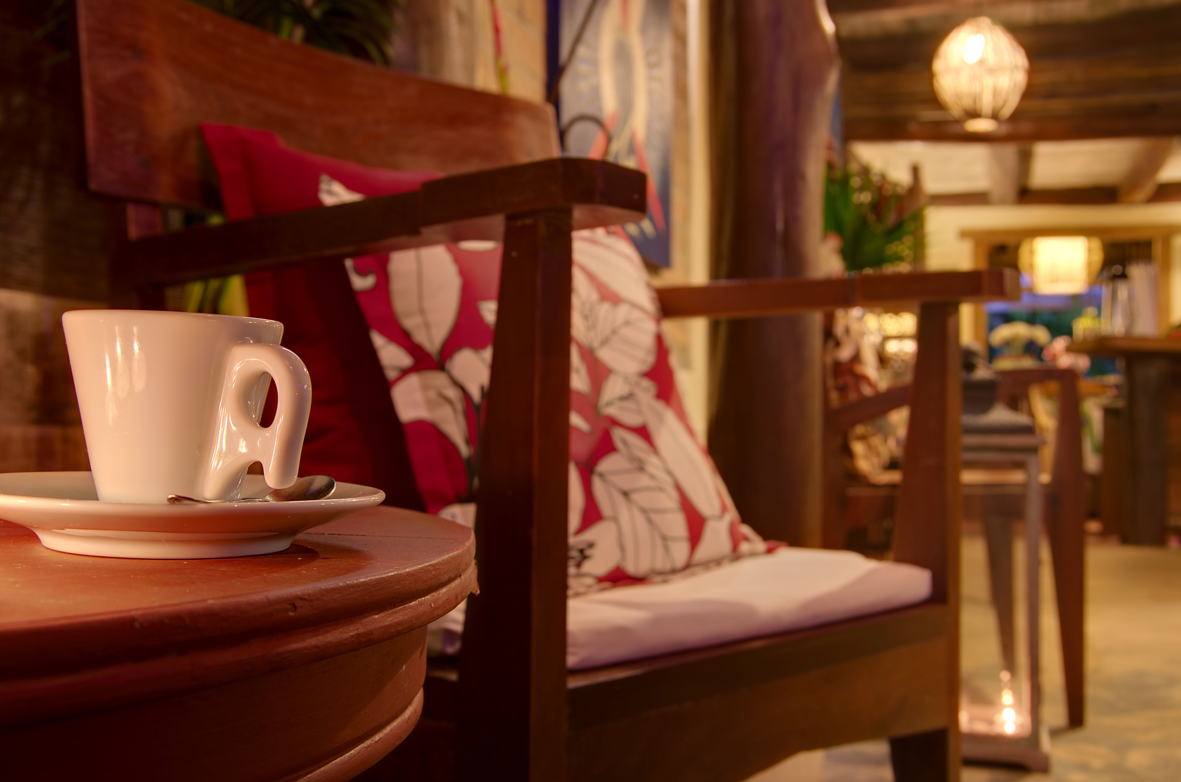 A white cup on a saucer on a wooden table, a wooden chair with a floral cushion, and a hanging light fixture in a warmly lit room.