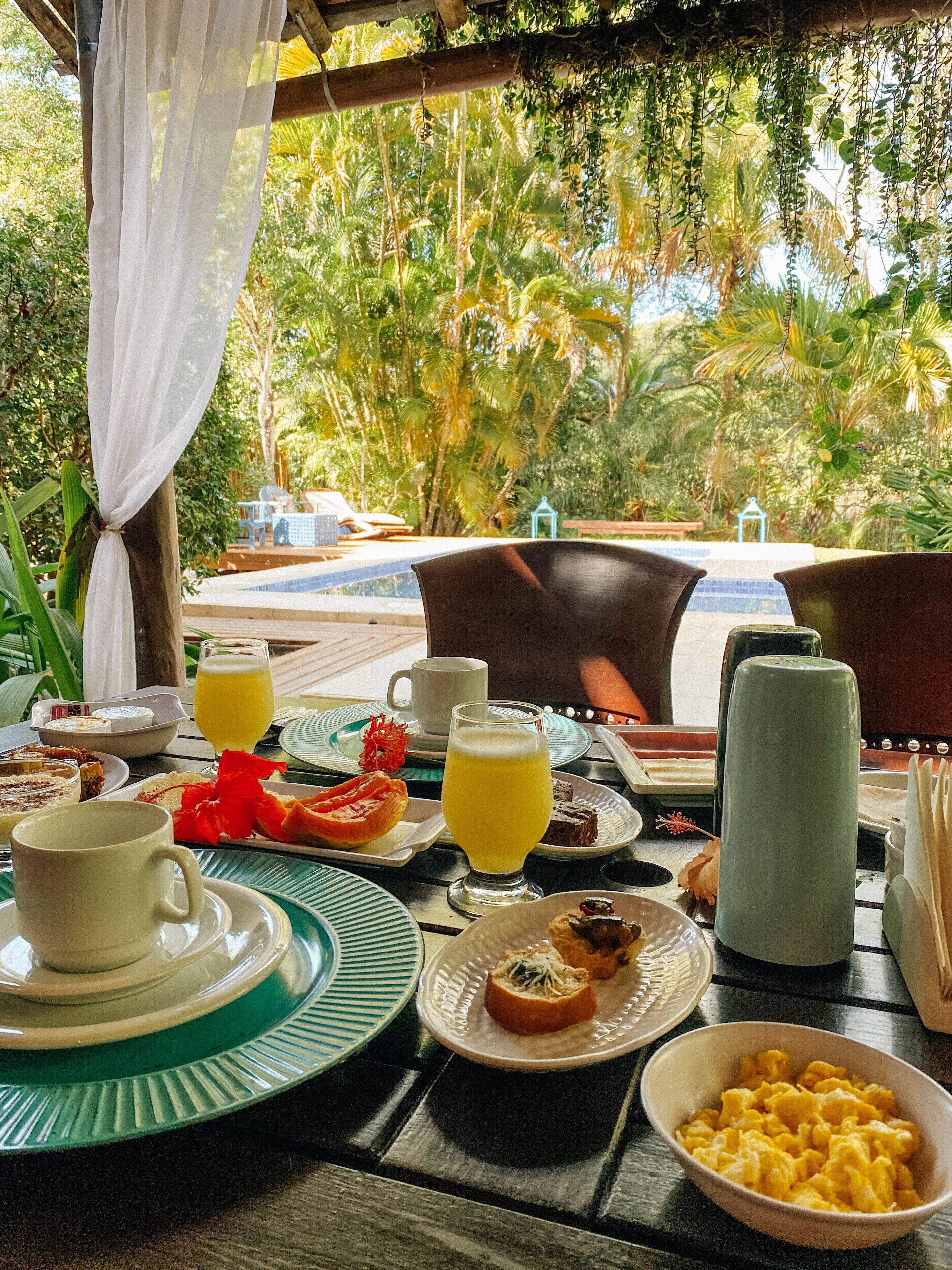 A breakfast table set with plates, cups, and a bowl of scrambled eggs, overlooking a pool and lush greenery.