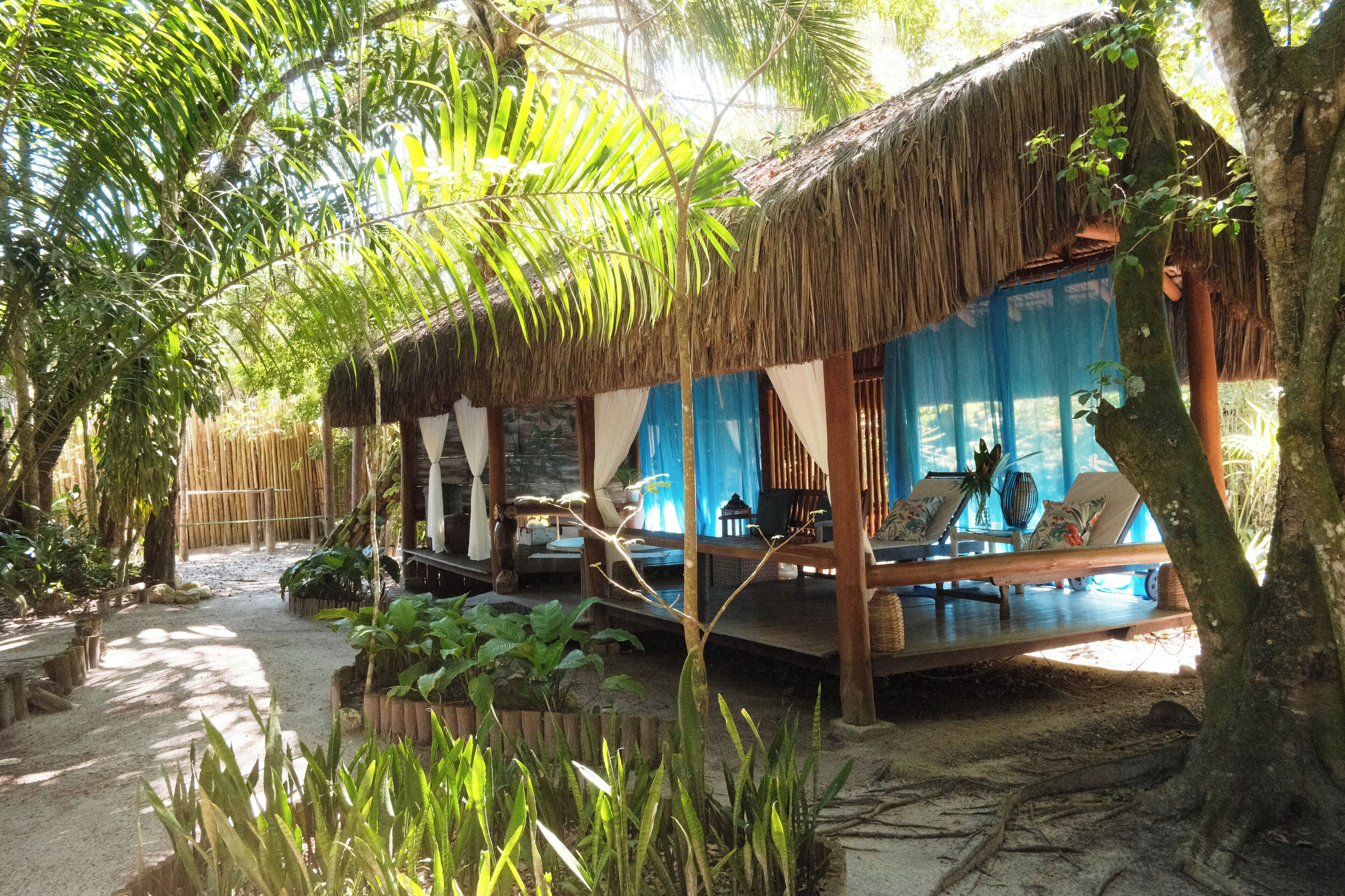 A thatched-roof structure with a covered seating area, surrounded by lush greenery and a sandy path.