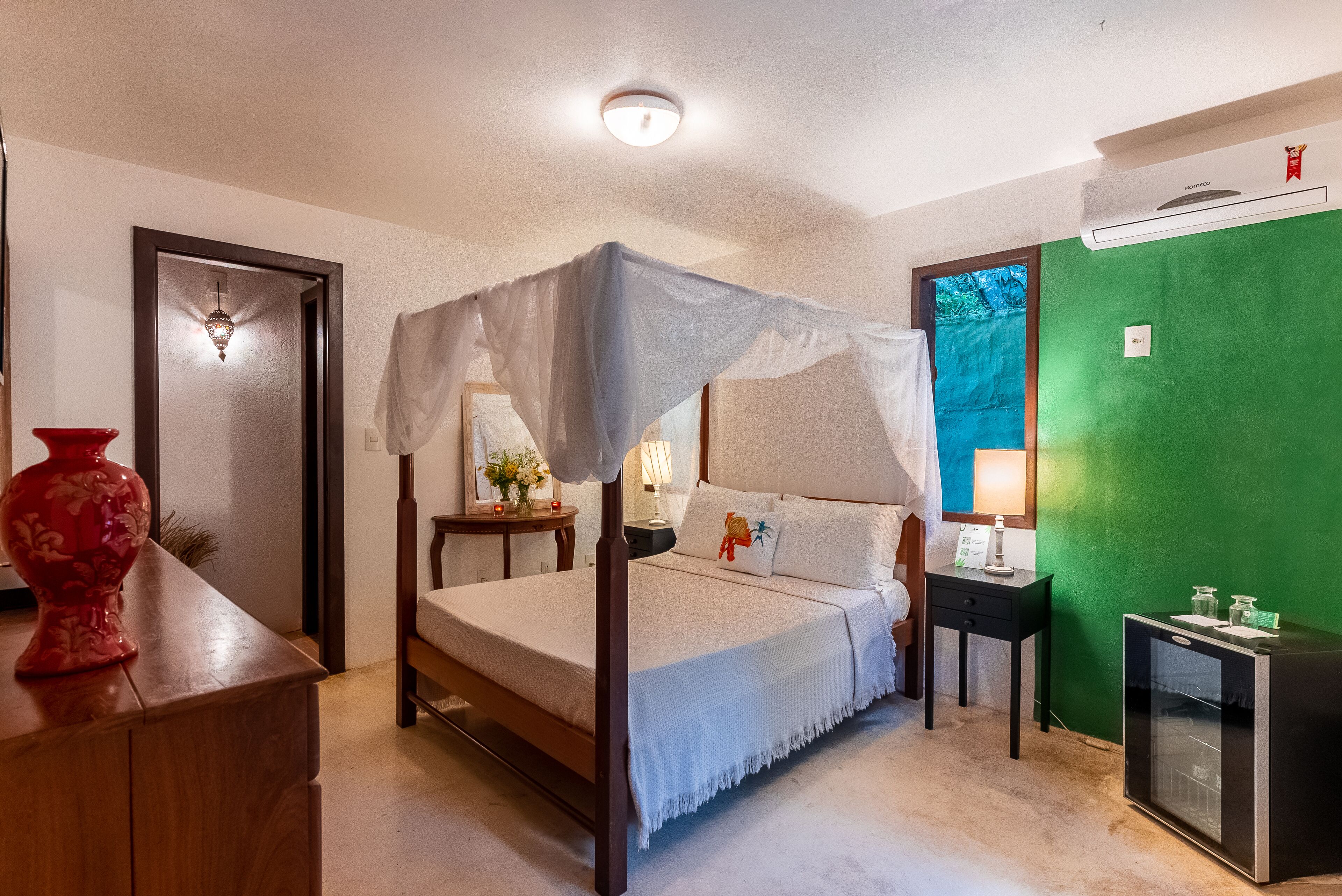 A room with a four-poster bed, a small bedside table, a red vase, a green wall, and a mini-fridge.