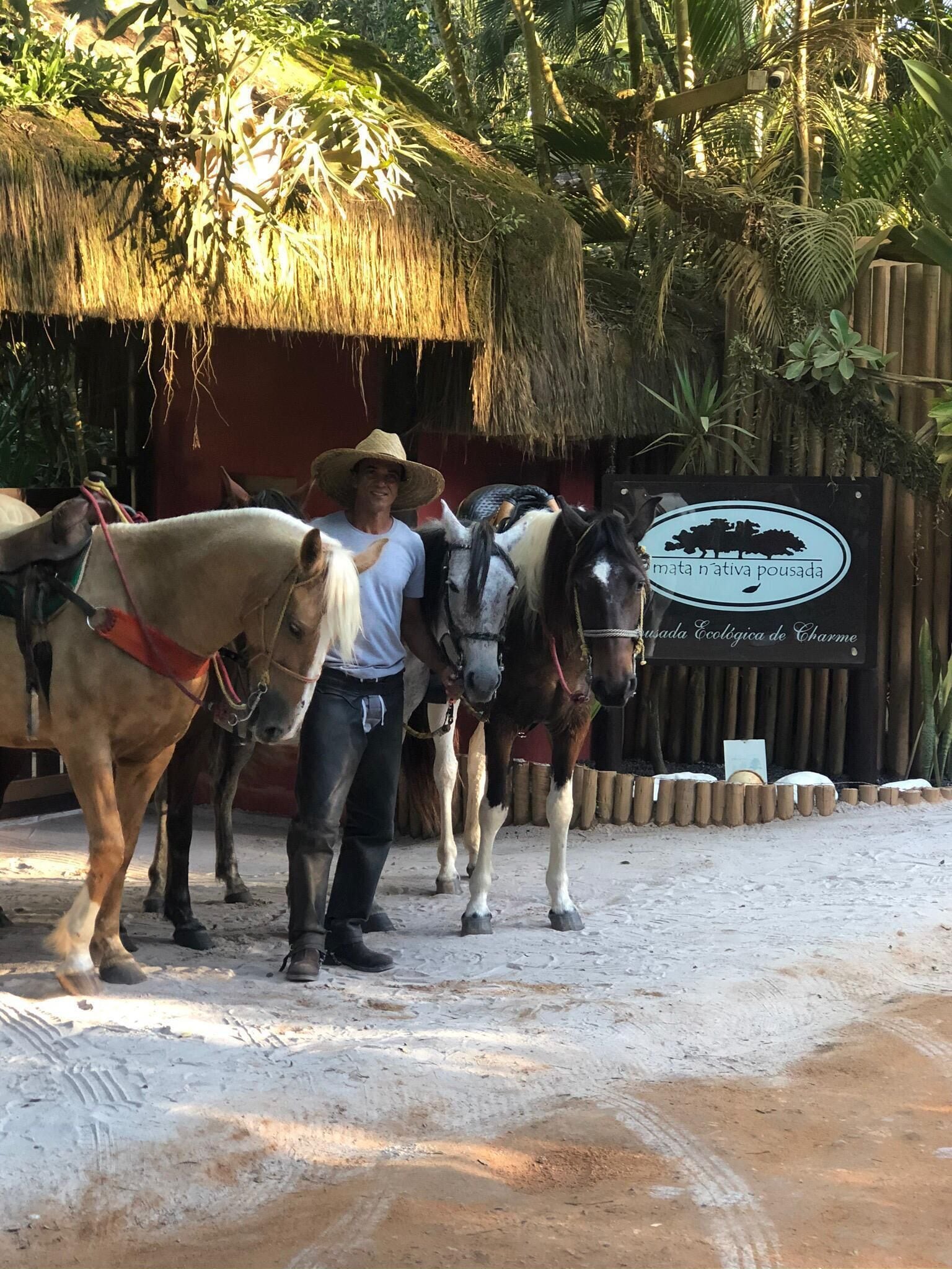 Un uomo in piedi accanto a due cavalli in un ambiente di stalla con tetto in paglia e un cartello che indica un centro di allevamento di cavalli.