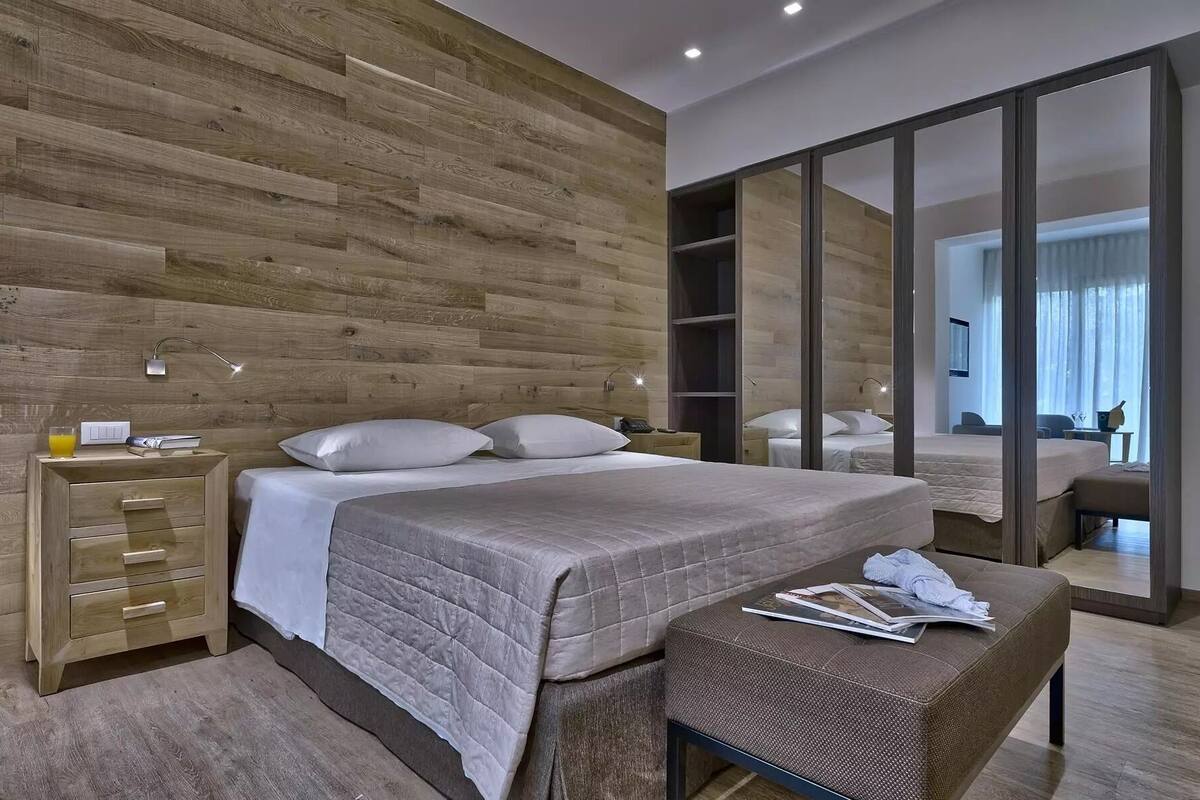 A modern bedroom with a wooden wall, a large bed with a quilted cover, a bedside table with a lamp, and a mirror.
