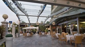 Un área de café al aire libre con mesas y sillas, estructura de techo de vidrio e iluminación decorativa.