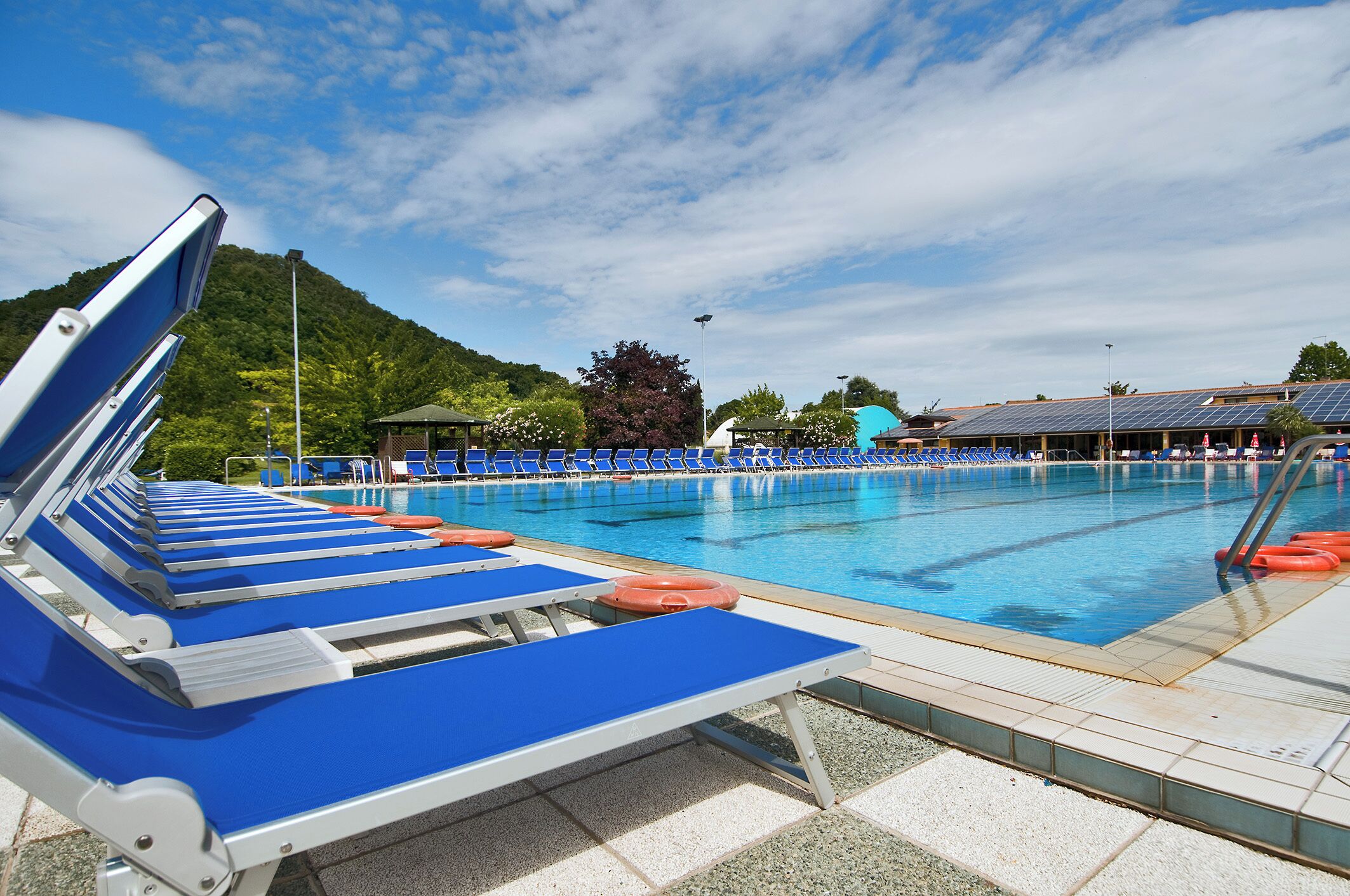 Una piscina con sillas reclinables azules y boyas salvavidas naranjas, rodeada de un solárium con baldosas y una montaña al fondo.