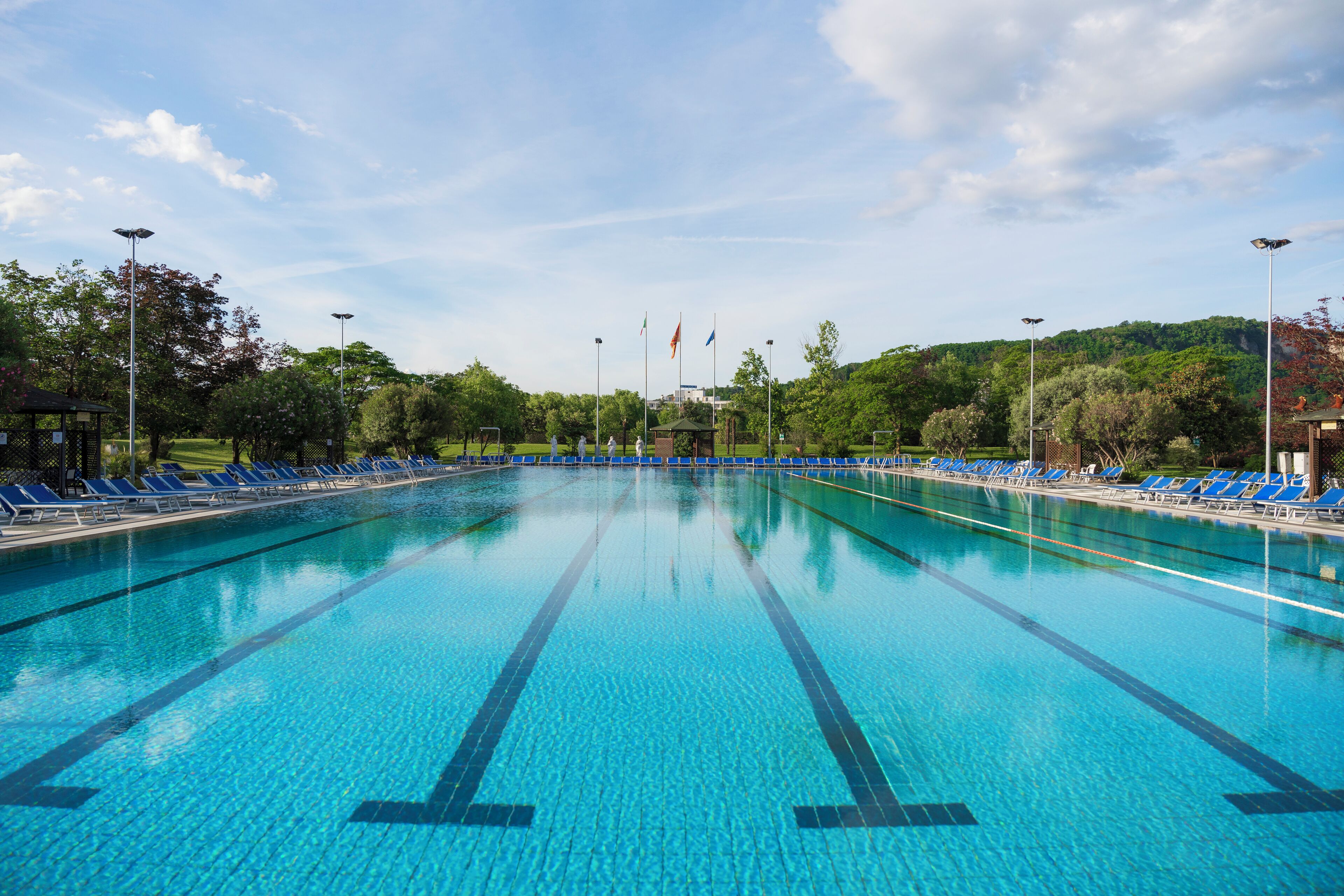A large, clear swimming pool with blue lane markings, surrounded by blue lounge chairs and trees.