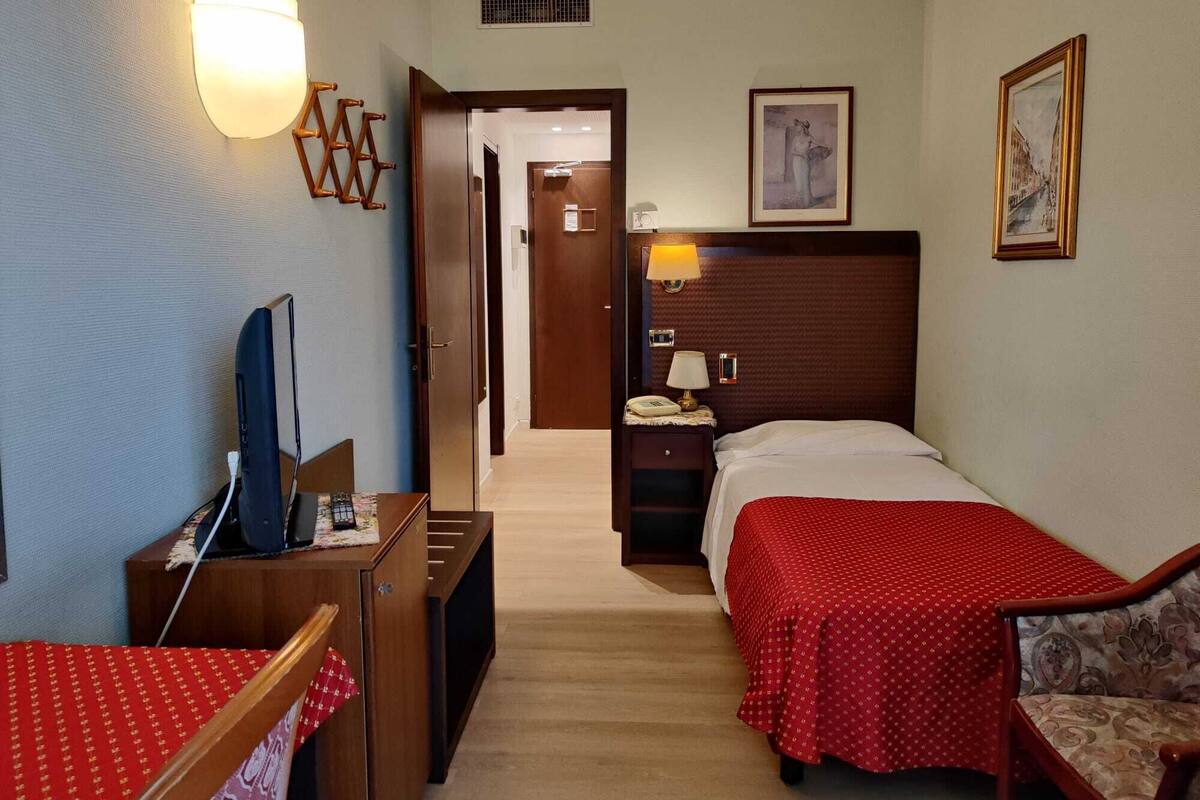 A hotel room with a bed, a desk with a TV, a chair, a bedside table with a lamp, and a framed picture on the wall.