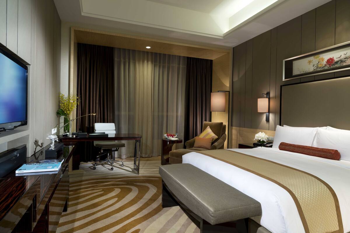 A hotel room with a large bed, a desk with a computer, a chair, a television, and a decorative plant.