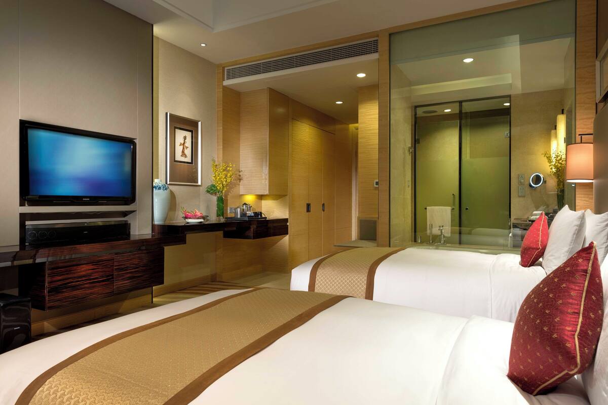 A hotel room with two beds, a TV, a minibar, and a bathroom visible through a glass door.