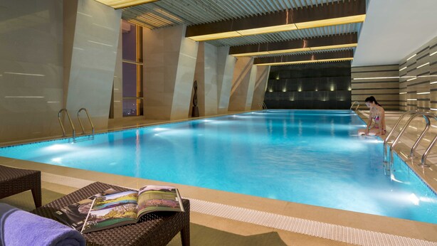 Une piscine intérieure à l’eau claire, un coin salon avec des magazines et une personne assise au bord de la piscine.