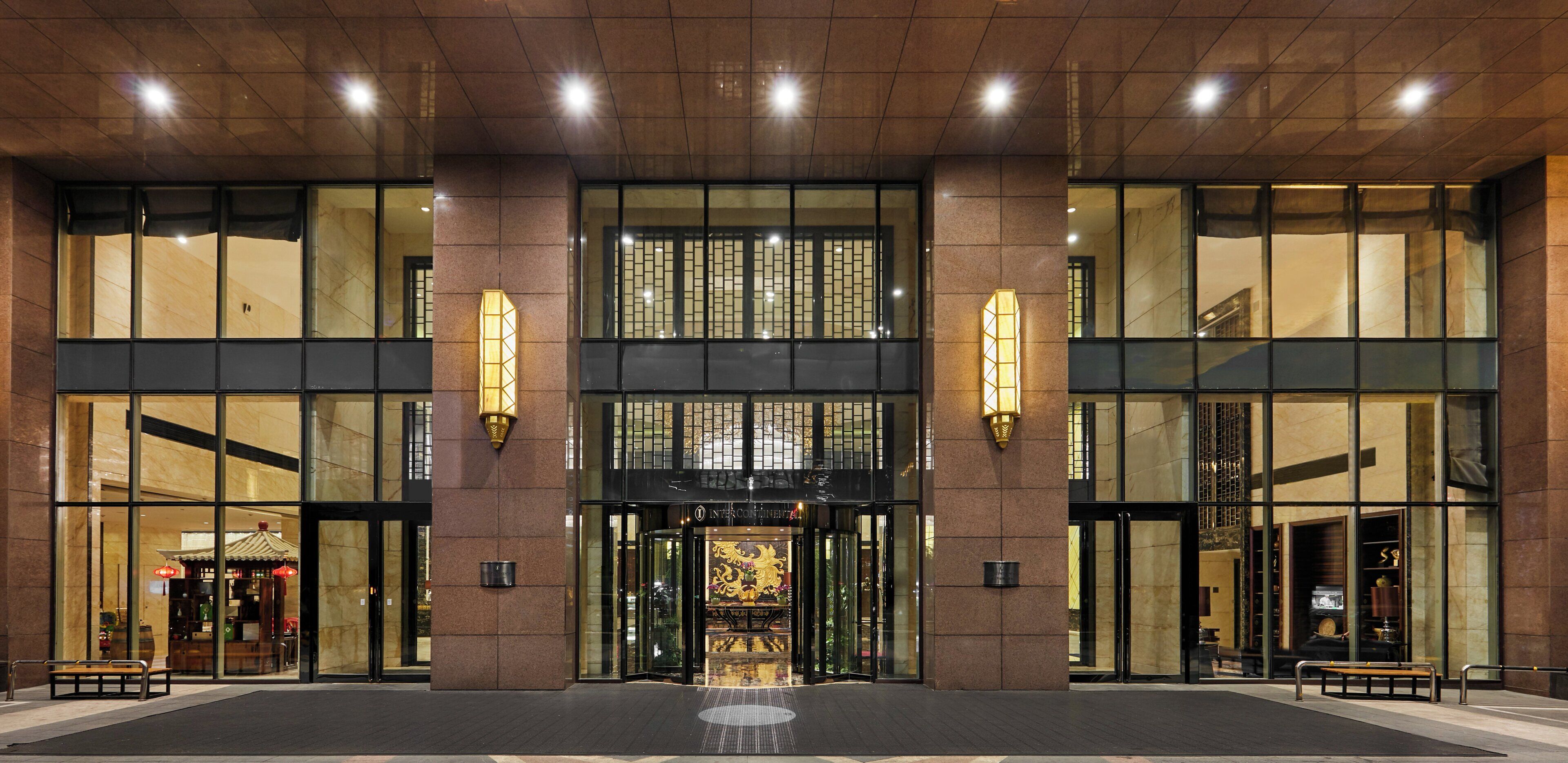 A hotel entrance with a glass facade, a traditional Asian-style gate, and a well-lit interior.